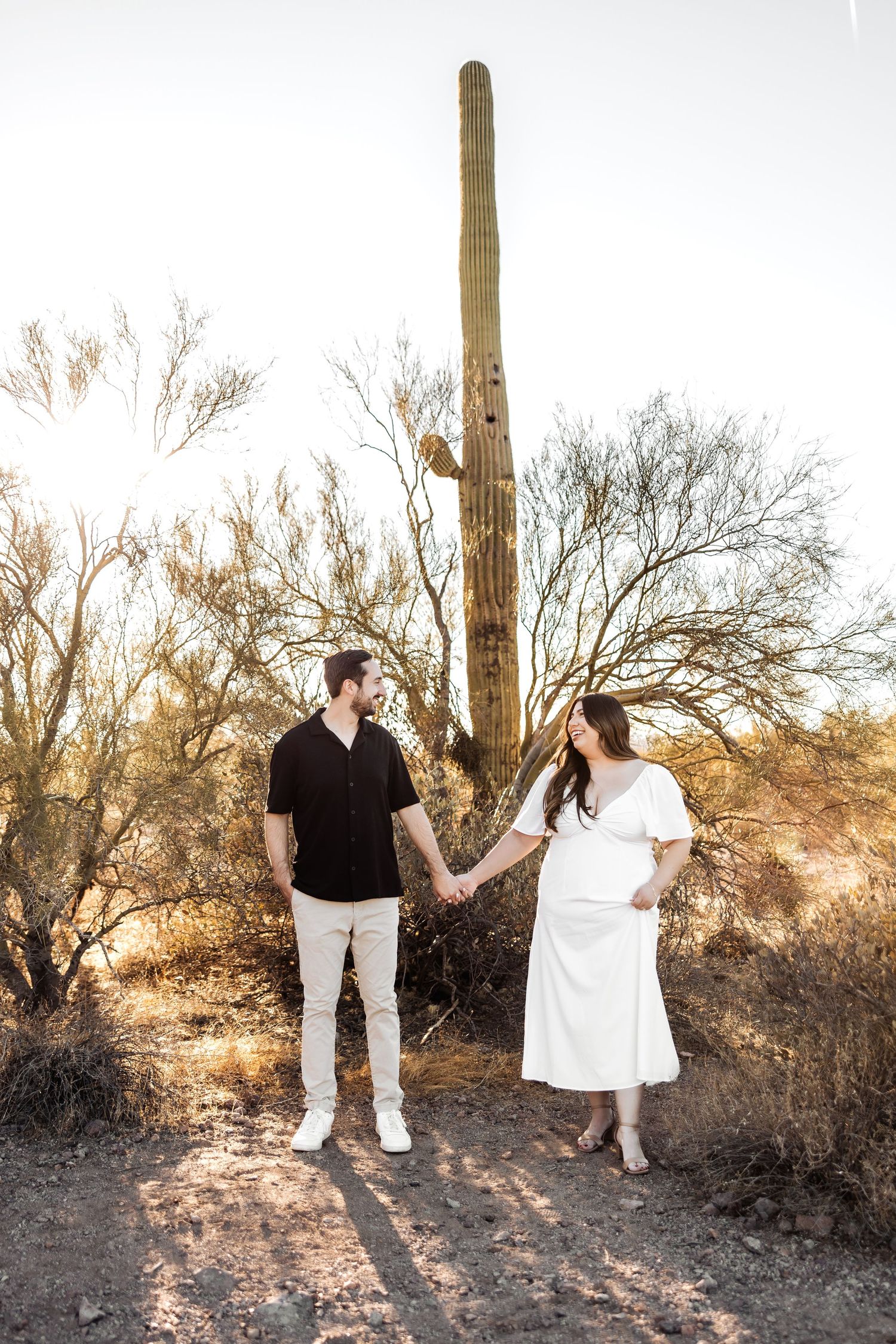 Couple holding hands in front of tall saguaro cactus during golden desert sunset in Arizona landscape.