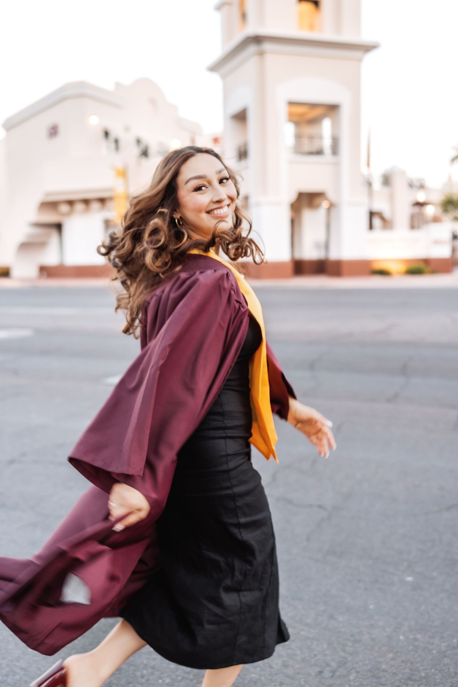 Graduate in flowing maroon gown and gold sash twirls joyfully in front of white mission-style building.