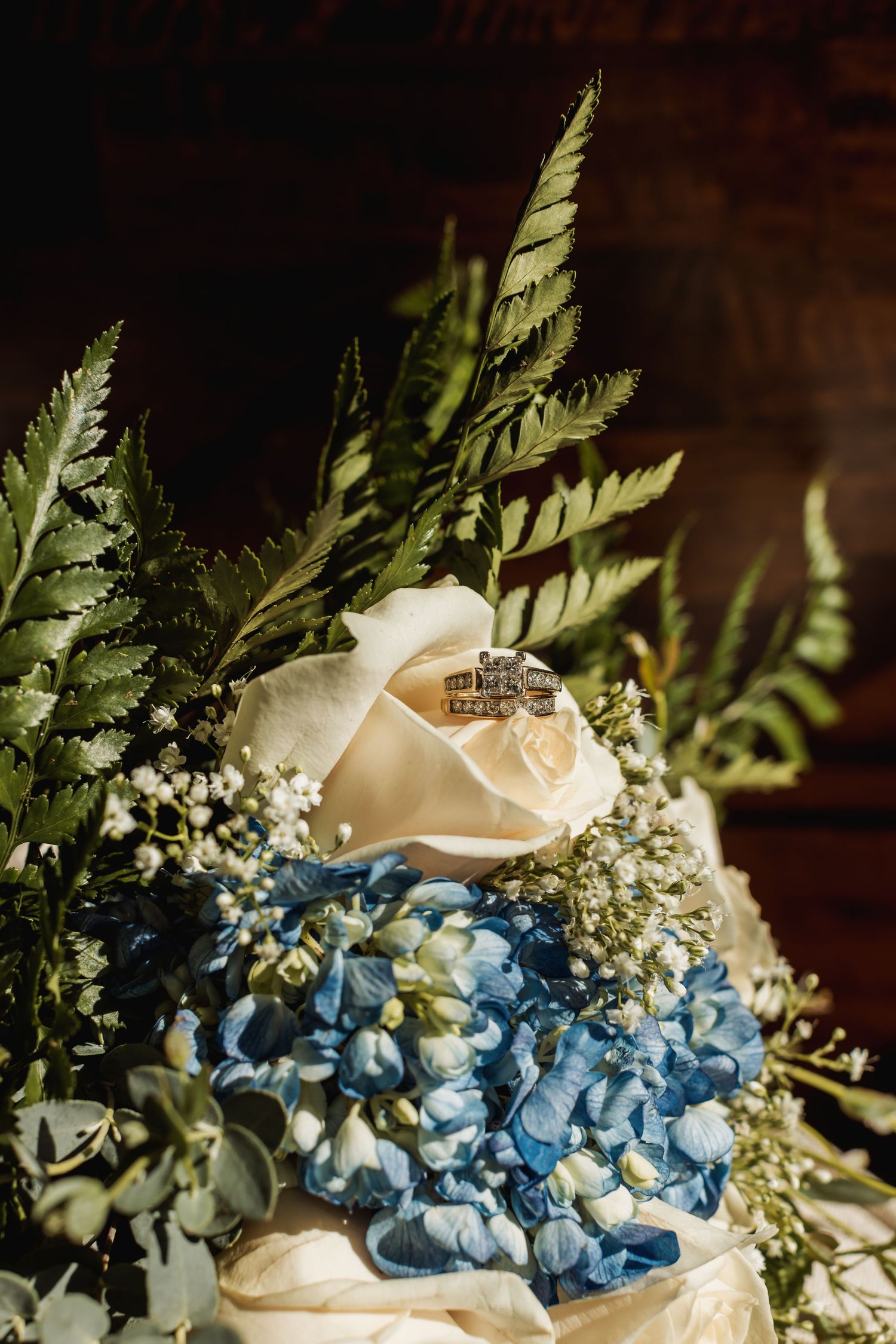 Wedding rings rest on a cream rose surrounded by blue hydrangeas and delicate ferns in an elegant floral arrangement.