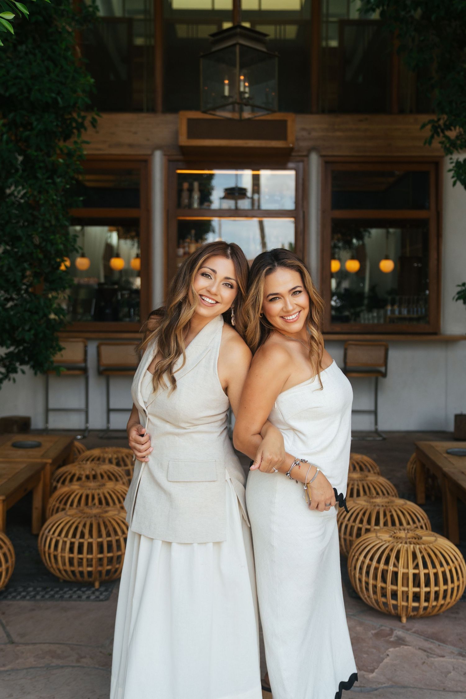 Two women in white dresses pose together in a space with rattan furniture and wooden floors.