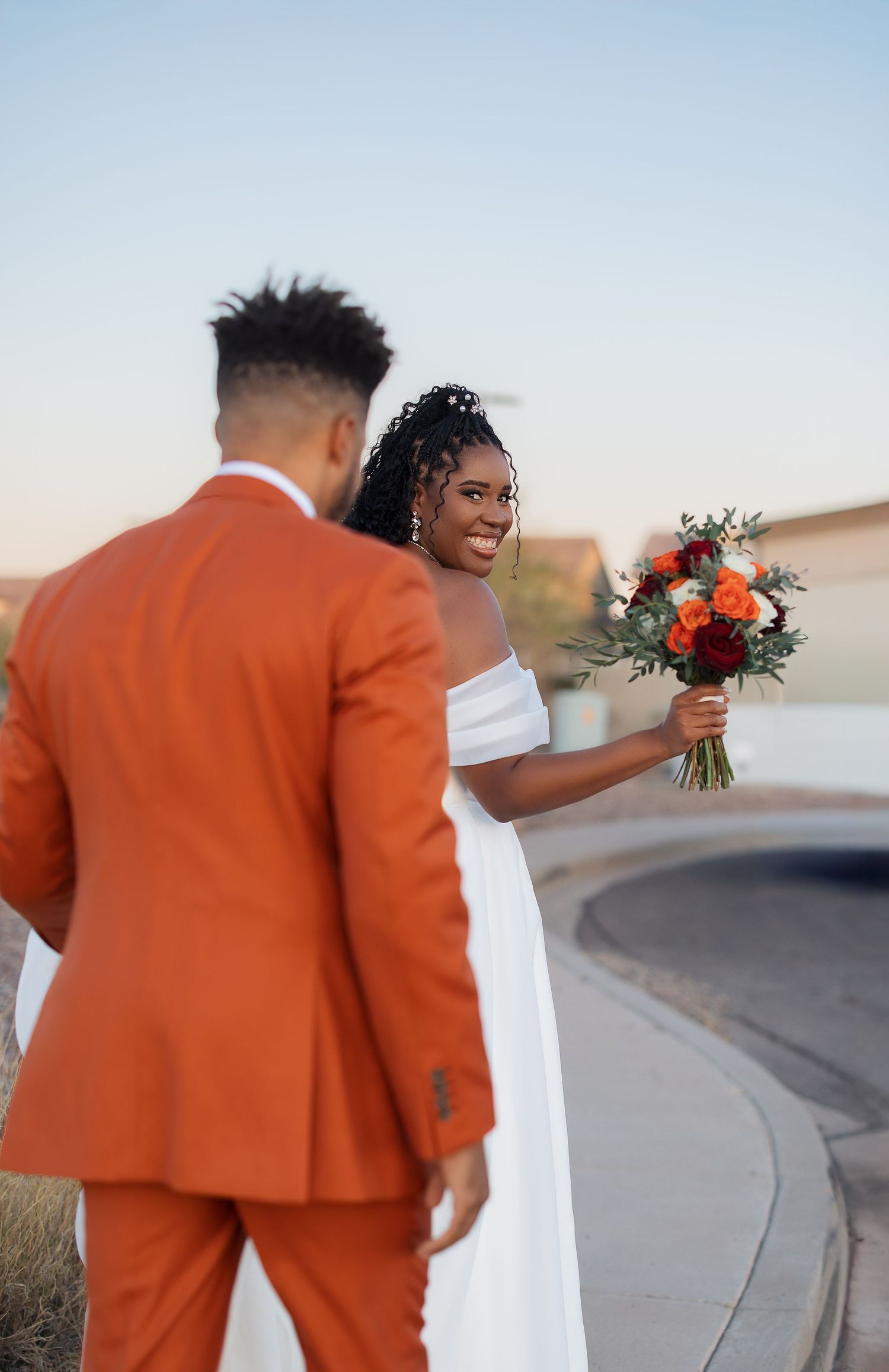 Couple walking along a curved desert road during sunset with orange and white wedding attire.