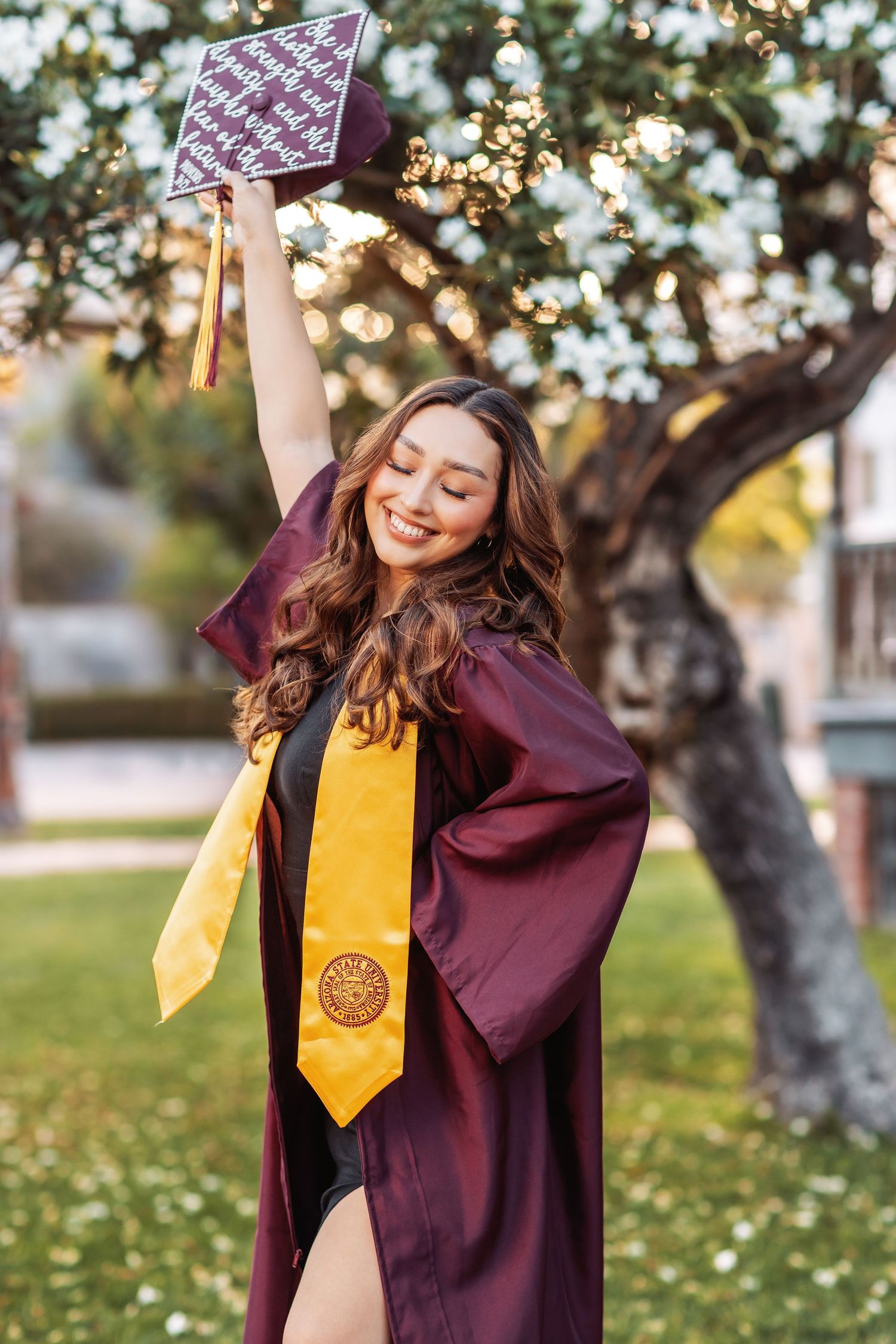 Graduate in maroon cap and gown with yellow stole celebrates with diploma outside on sunny day with bokeh lights.