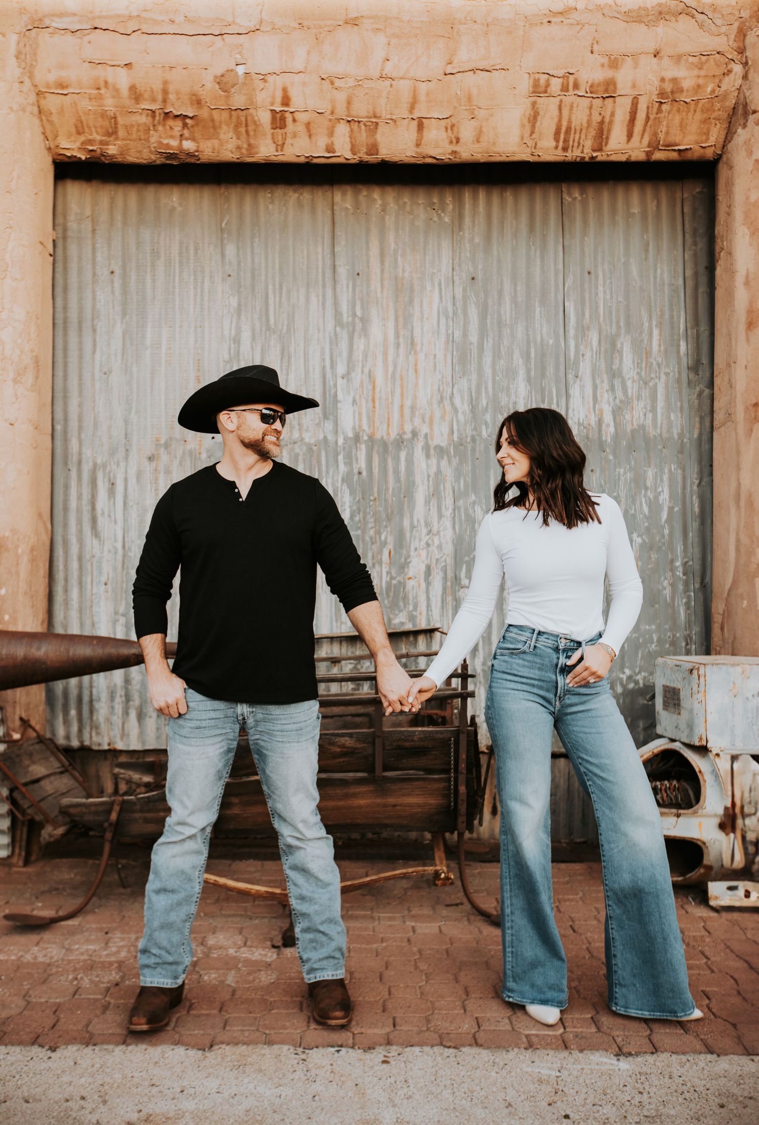 A couple in casual western wear holding hands against a rustic industrial door, wearing denim and contrasting tops.