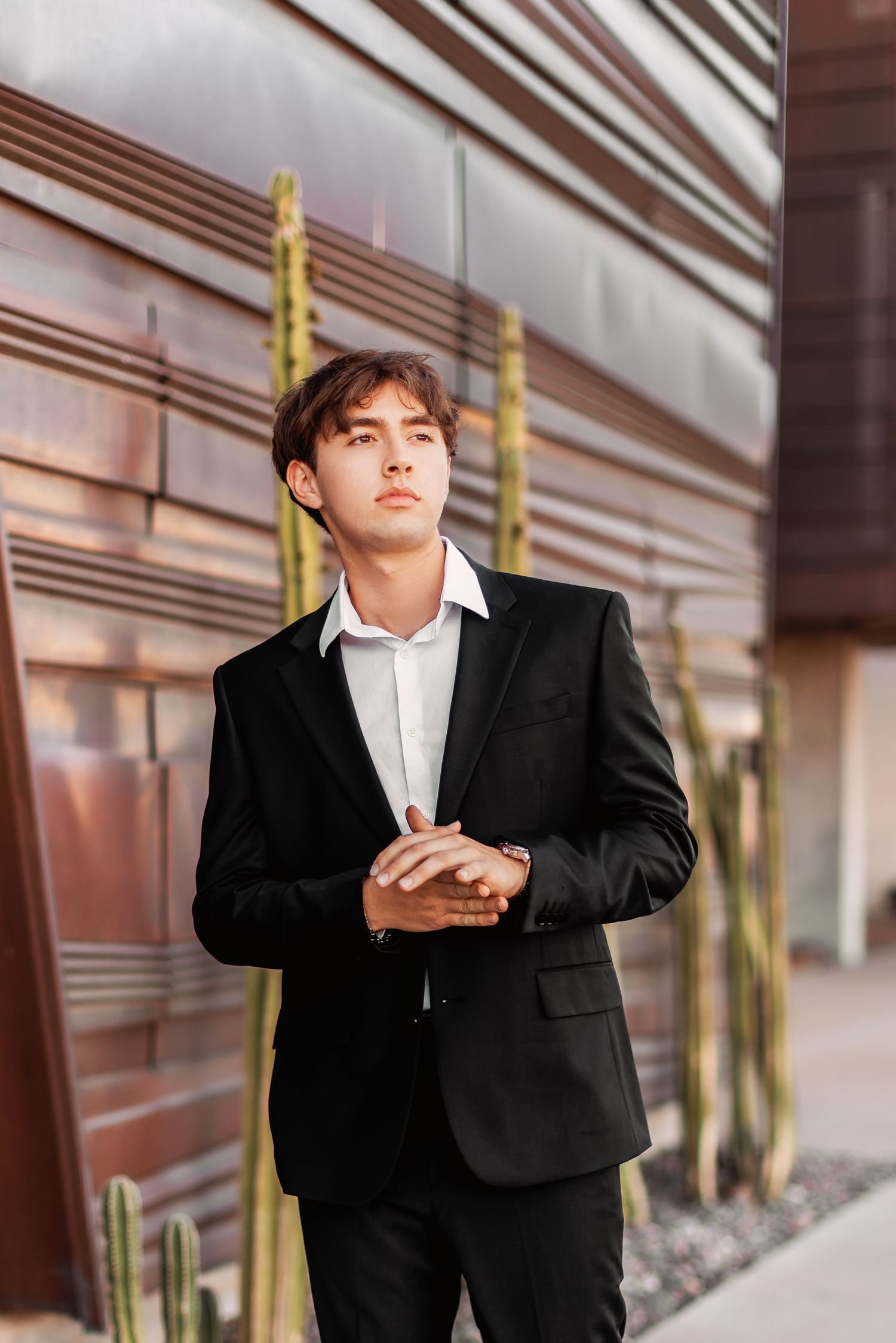 Professional portrait photo of someone in a black suit and white shirt standing against a modern architectural background.