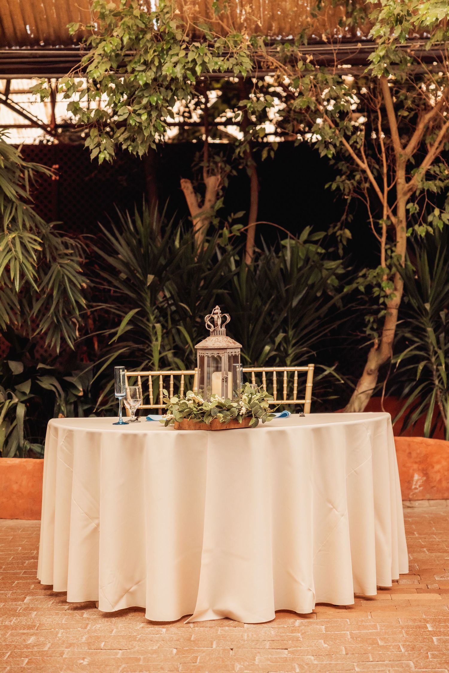 Round banquet tables with white tablecloths arranged on a brick patio with lush tropical garden backdrop and lantern decor.