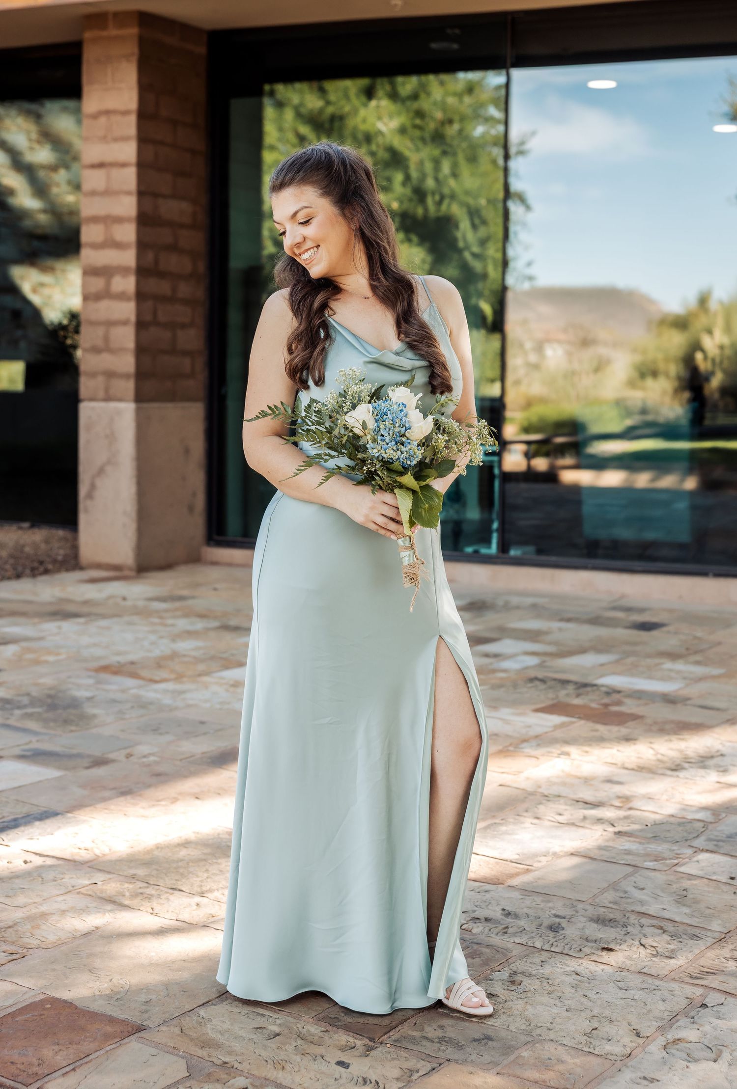 A person in a light mint green dress poses with a bouquet of flowers on a stone patio near brick pillars.