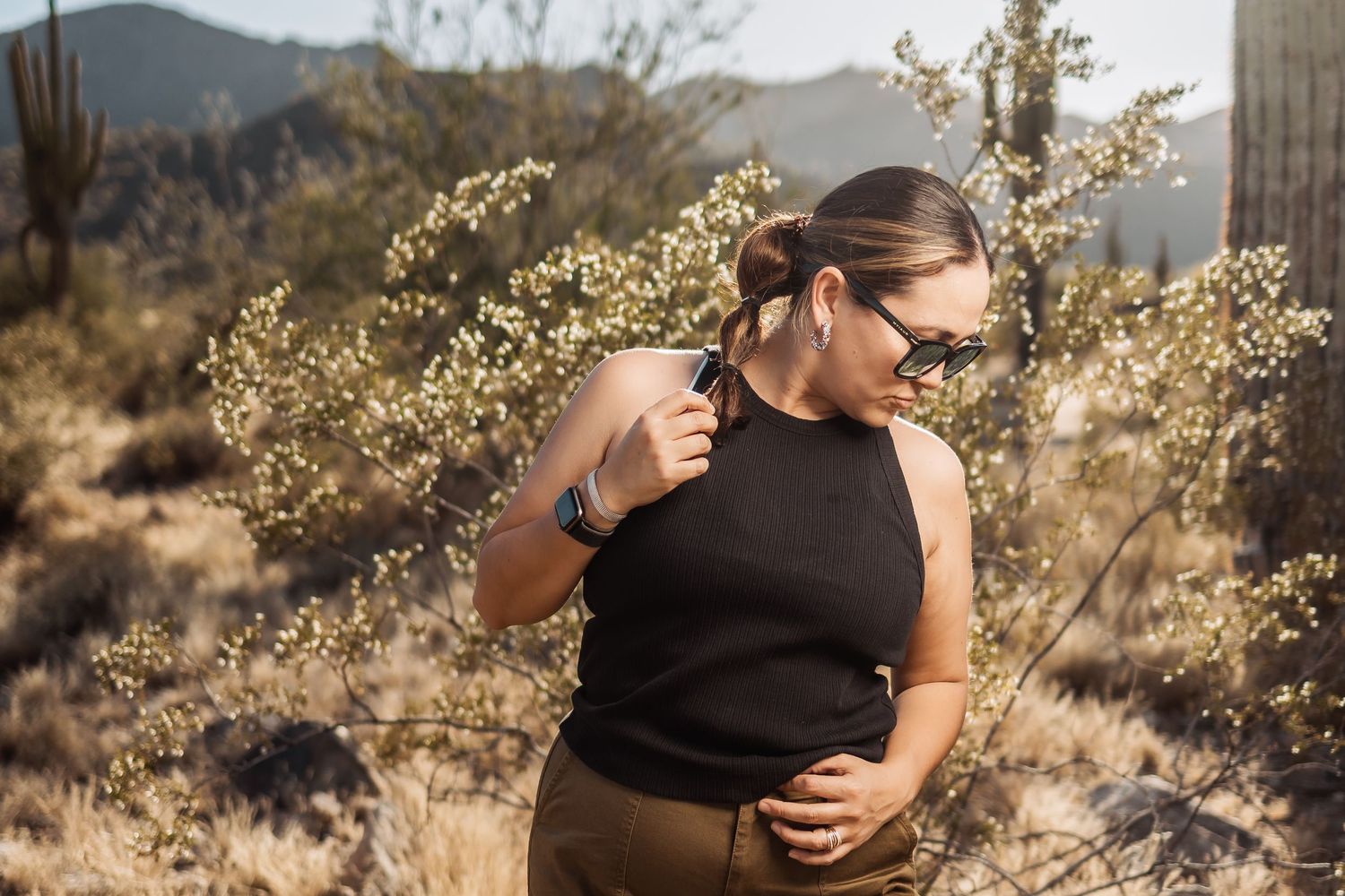 Someone in black sleeveless top poses by desert brush with sunlit mountains and desert flora in background.