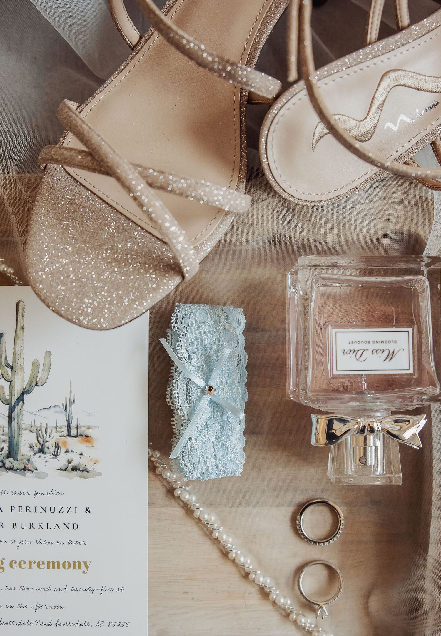 Wedding details flatlay with sparkly sandals, invitation with cactus design, perfume bottle, rings and lace garter on wooden surface.