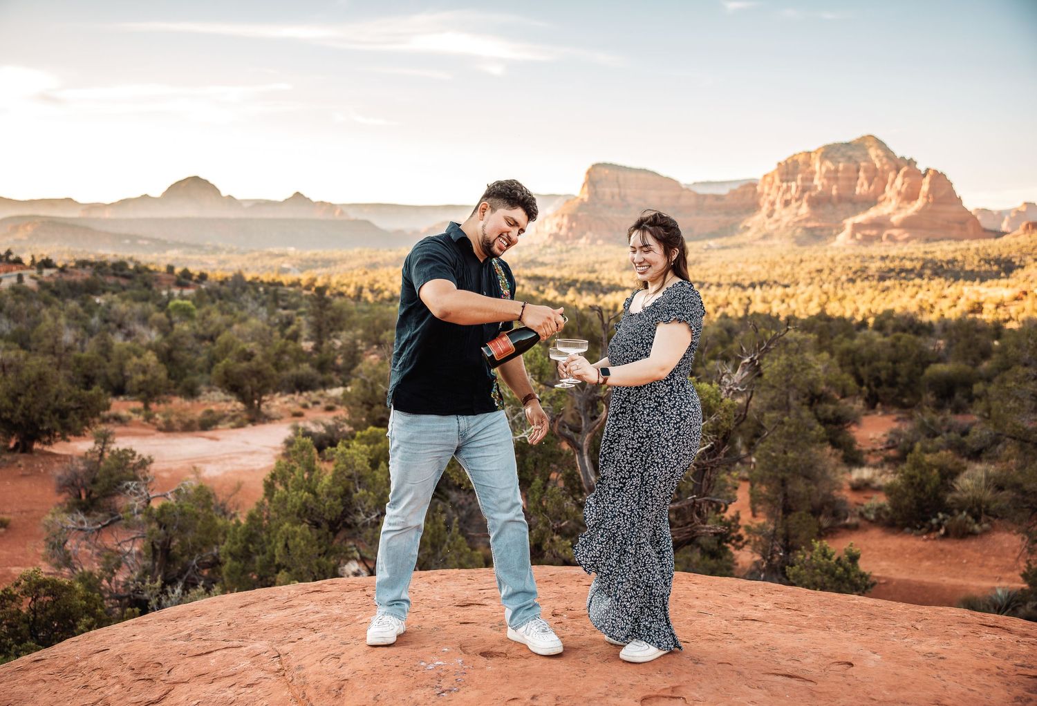 A couple shares a joyful moment together on red rocks overlooking scenic desert mountains during sunset.
