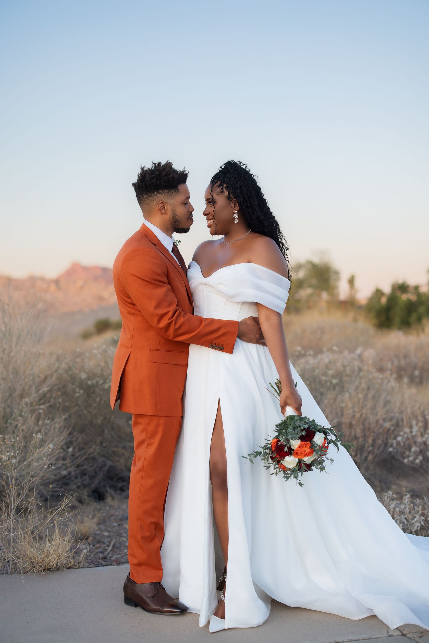 Newlywed couple embraces in white wedding dress and burnt orange suit against desert mountain backdrop at sunset.