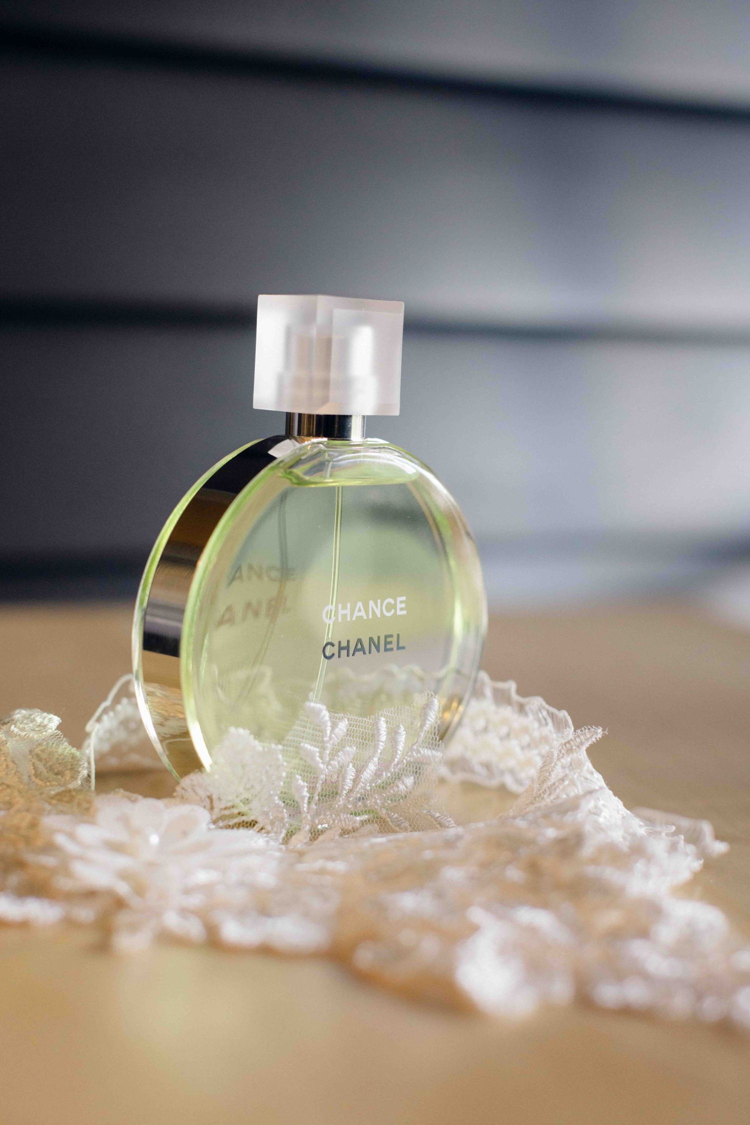 A Chanel Chance perfume bottle lies on a surface surrounded by delicate white lace fabric in a soft focus shot.