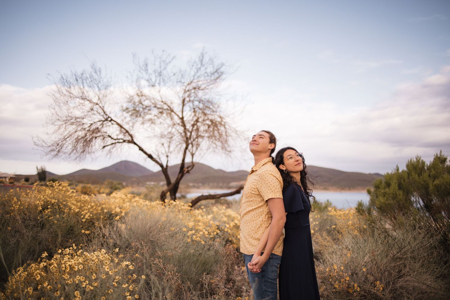 Couple stands back to back in a desert landscape with mountains and a lake in the background at sunset.