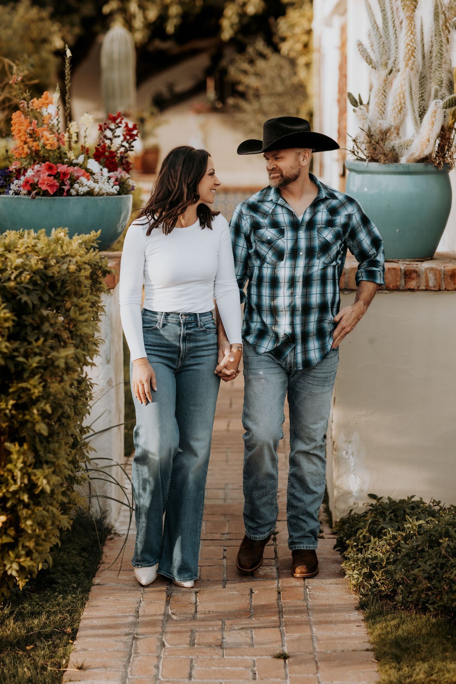 Couple in Western attire strolling along brick pathway surrounded by potted plants and autumn foliage.
