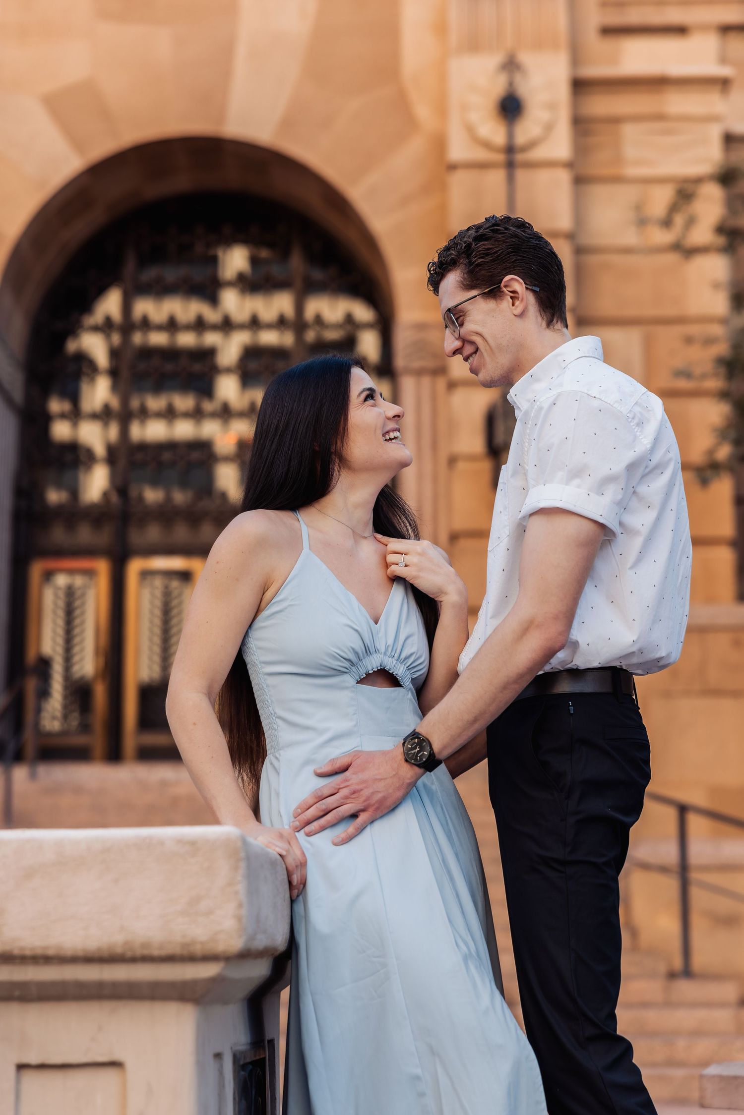 A couple embraces and shares a romantic moment in front of an ornate building with an arched doorway at sunset.