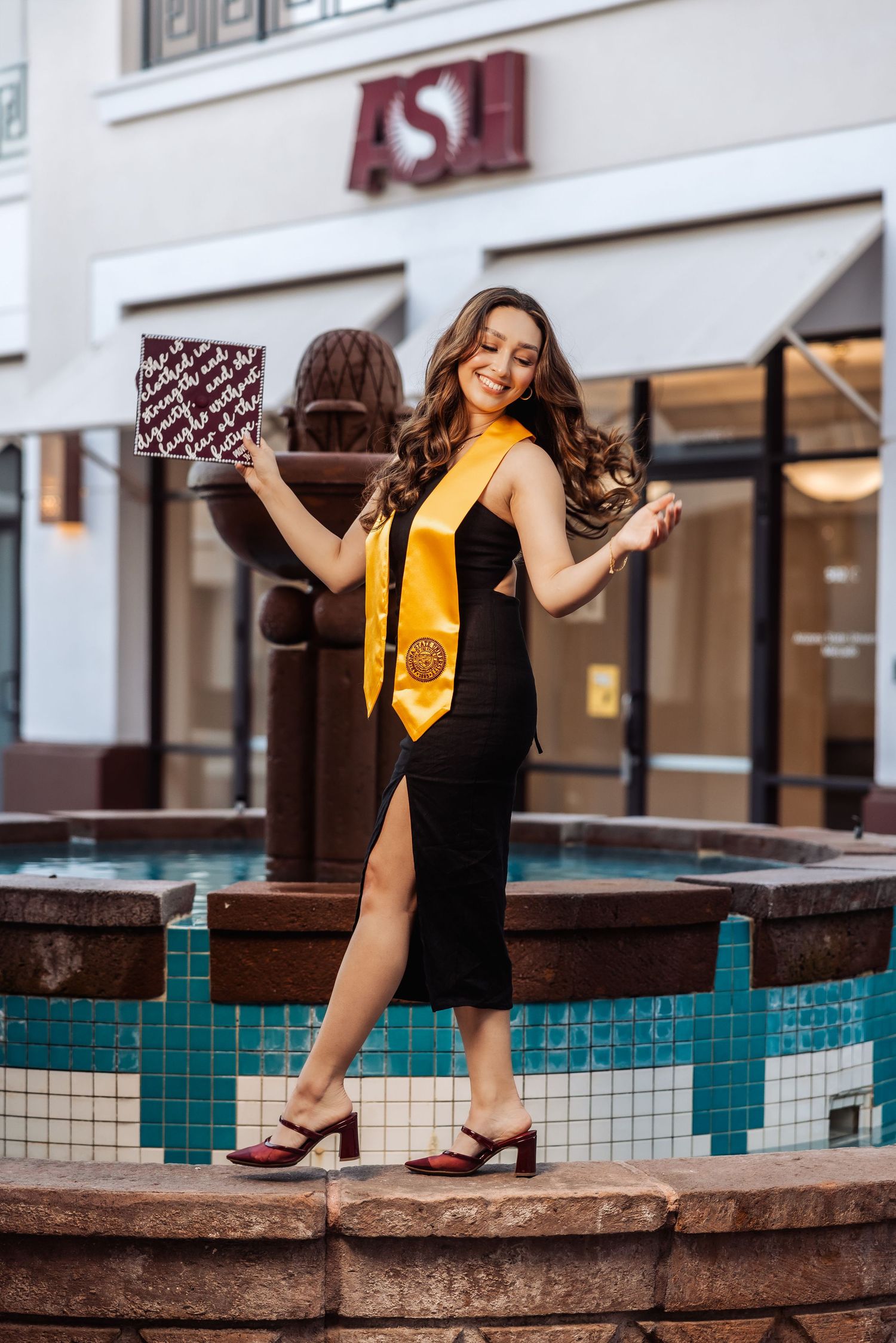 A graduate in a black dress and gold sash poses by a tiled fountain near an ASU storefront.