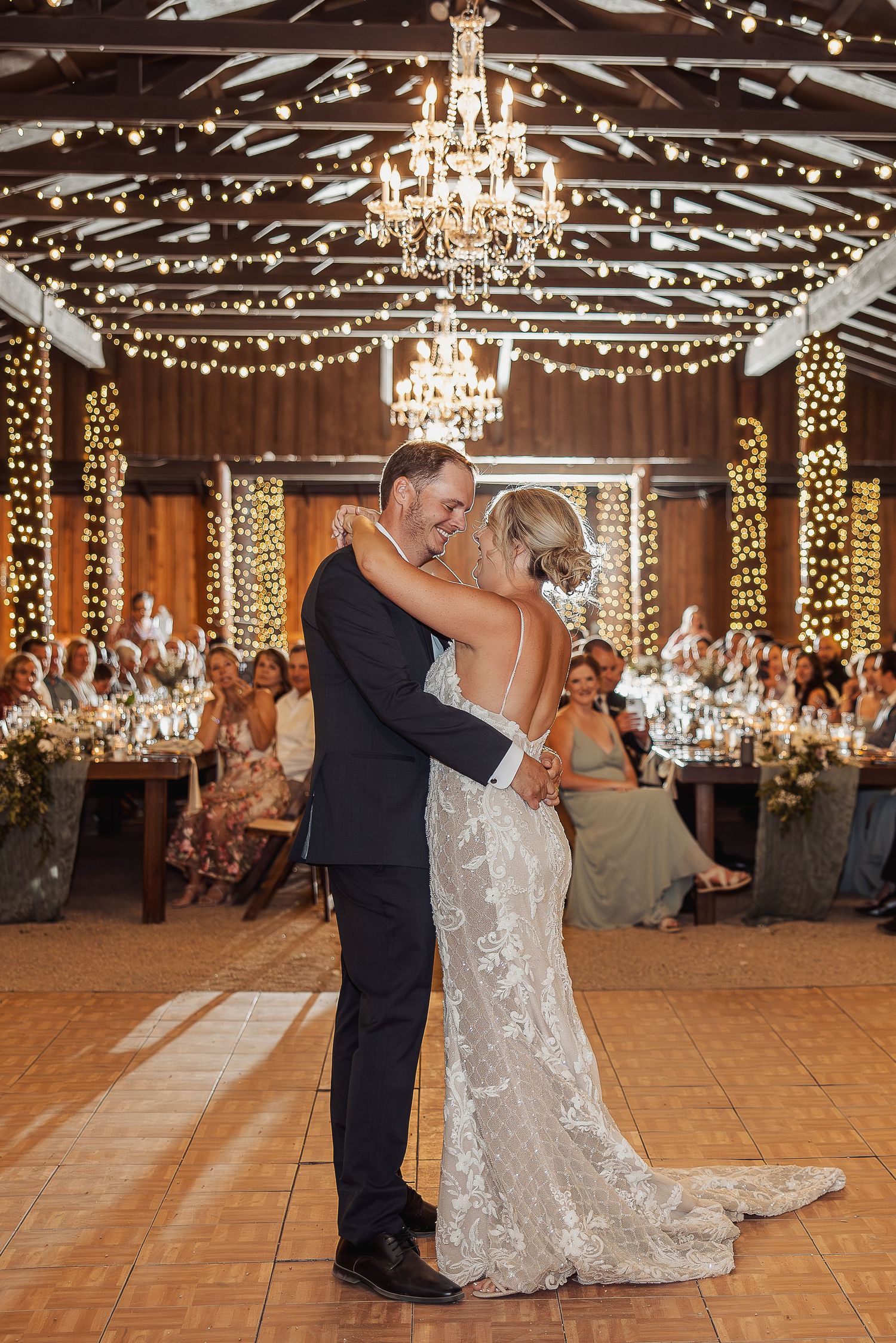 A couple shares their first dance in a rustic barn venue decorated with twinkling lights and elegant chandeliers.