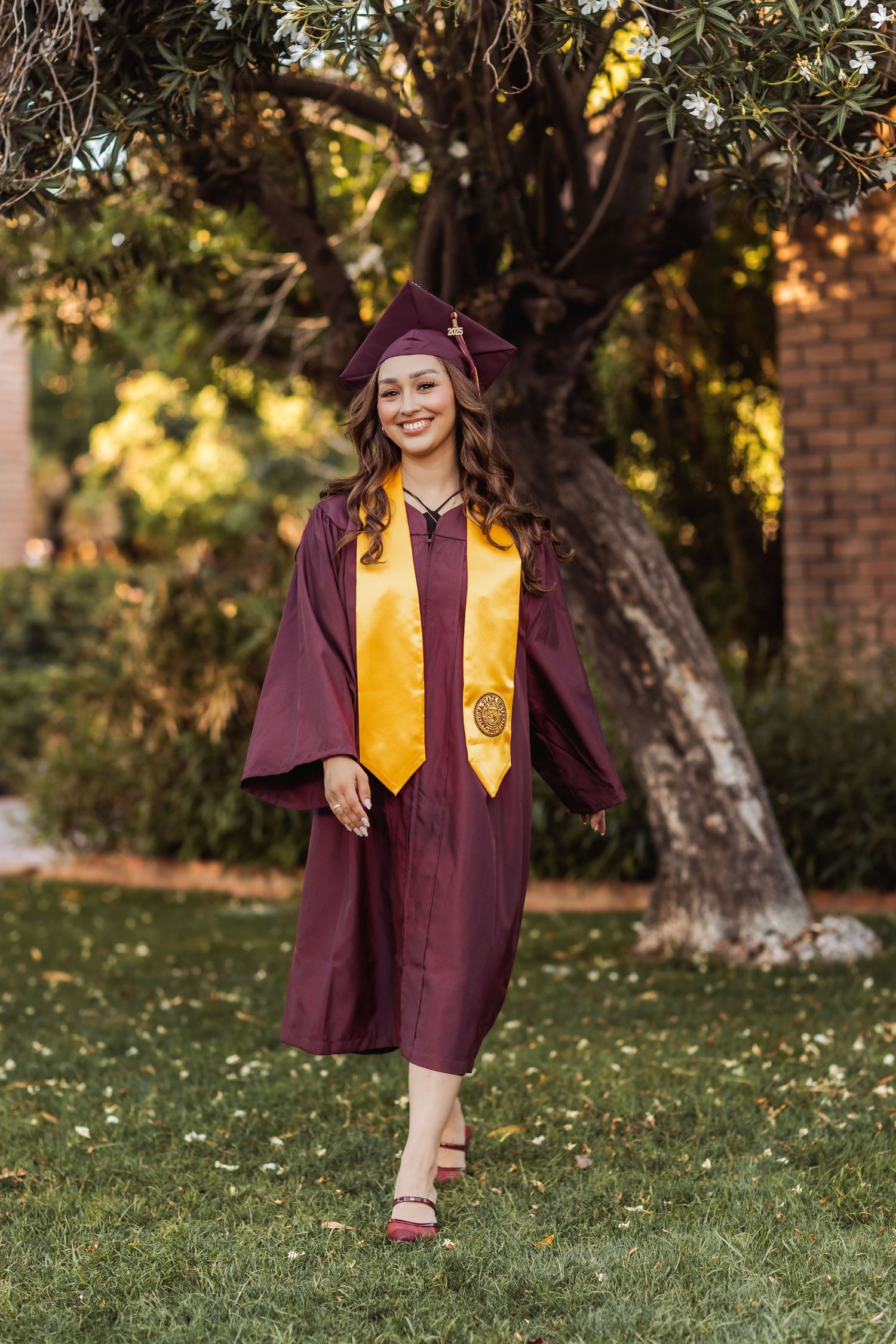 Graduate in maroon cap and gown with gold sash walks on grass during outdoor graduation photo session.