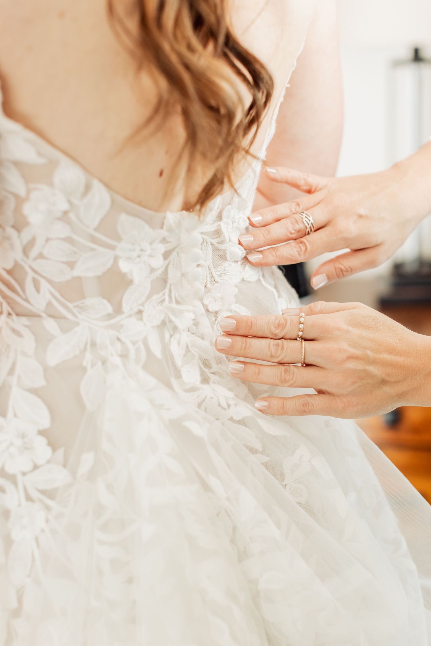 Close-up detail of delicate lace and beading on a white wedding dress with hands showing rings.