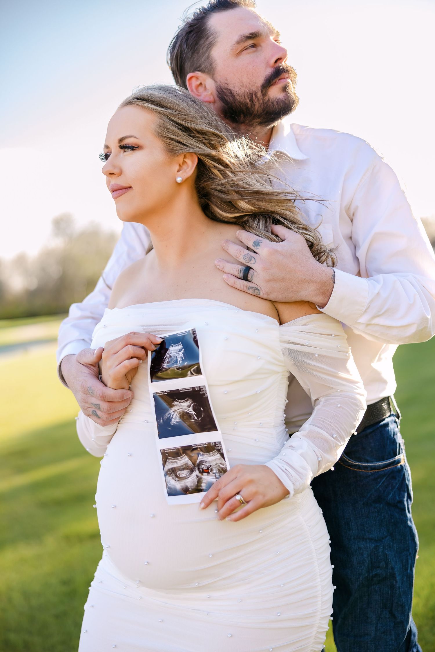 Couple in white and denim embrace while holding ultrasound photos during a pregnancy announcement photoshoot at sunset.