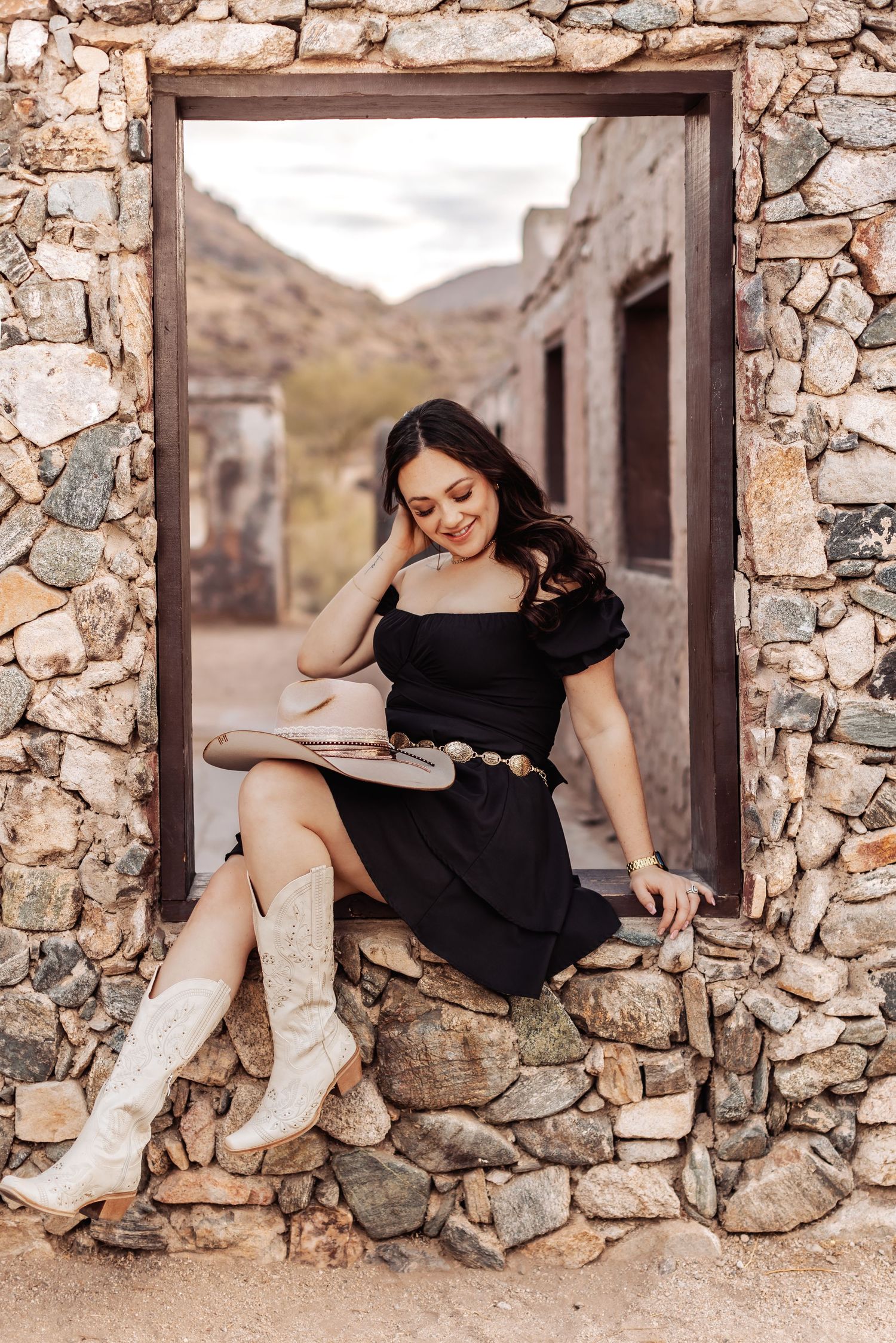 Woman in black dress and cowboy boots sits in rustic stone doorway reading a book in desert setting.