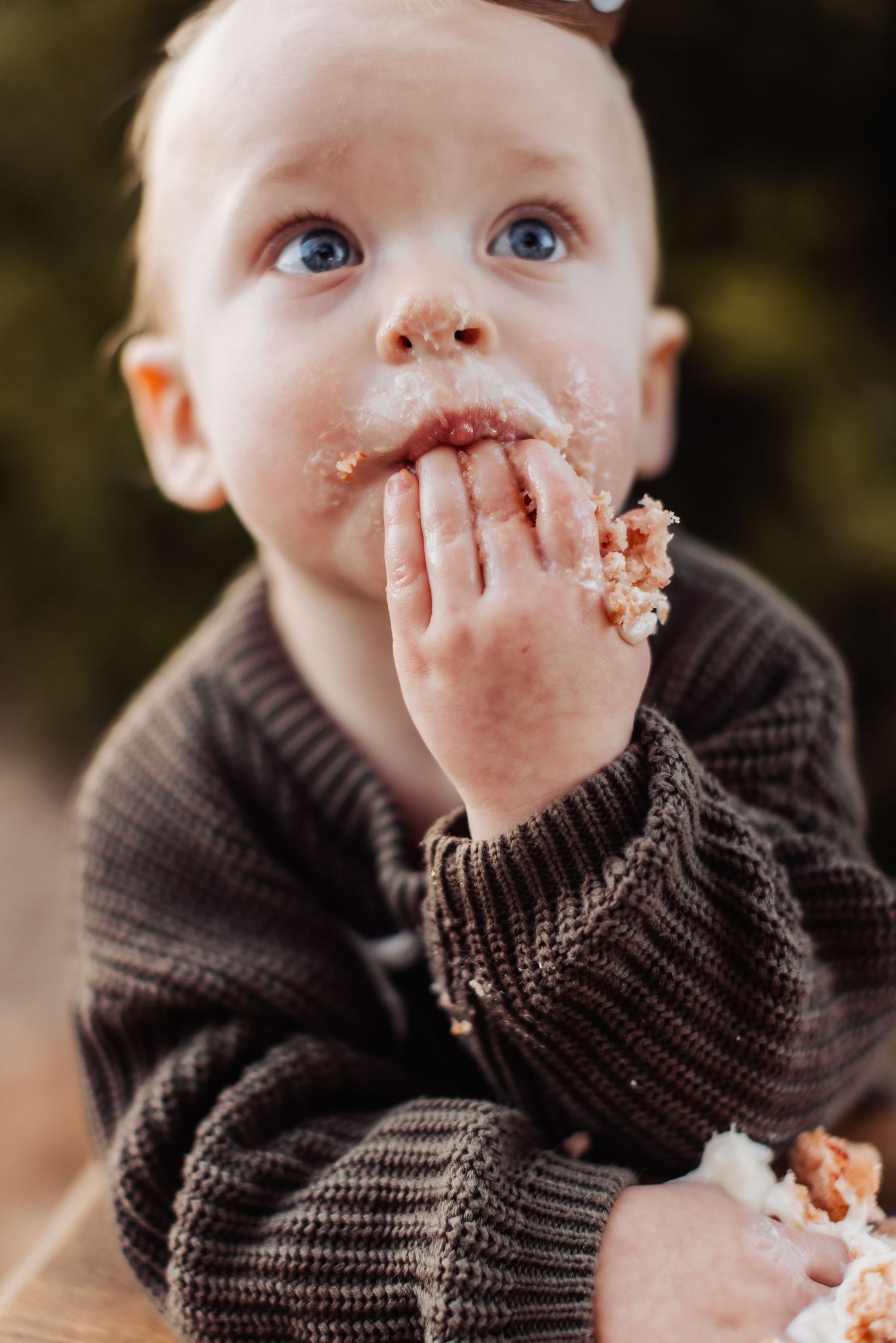 Baby wearing brown knit sweater messily eating cake with frosting on hands and face and bright blue eyes looking up.
