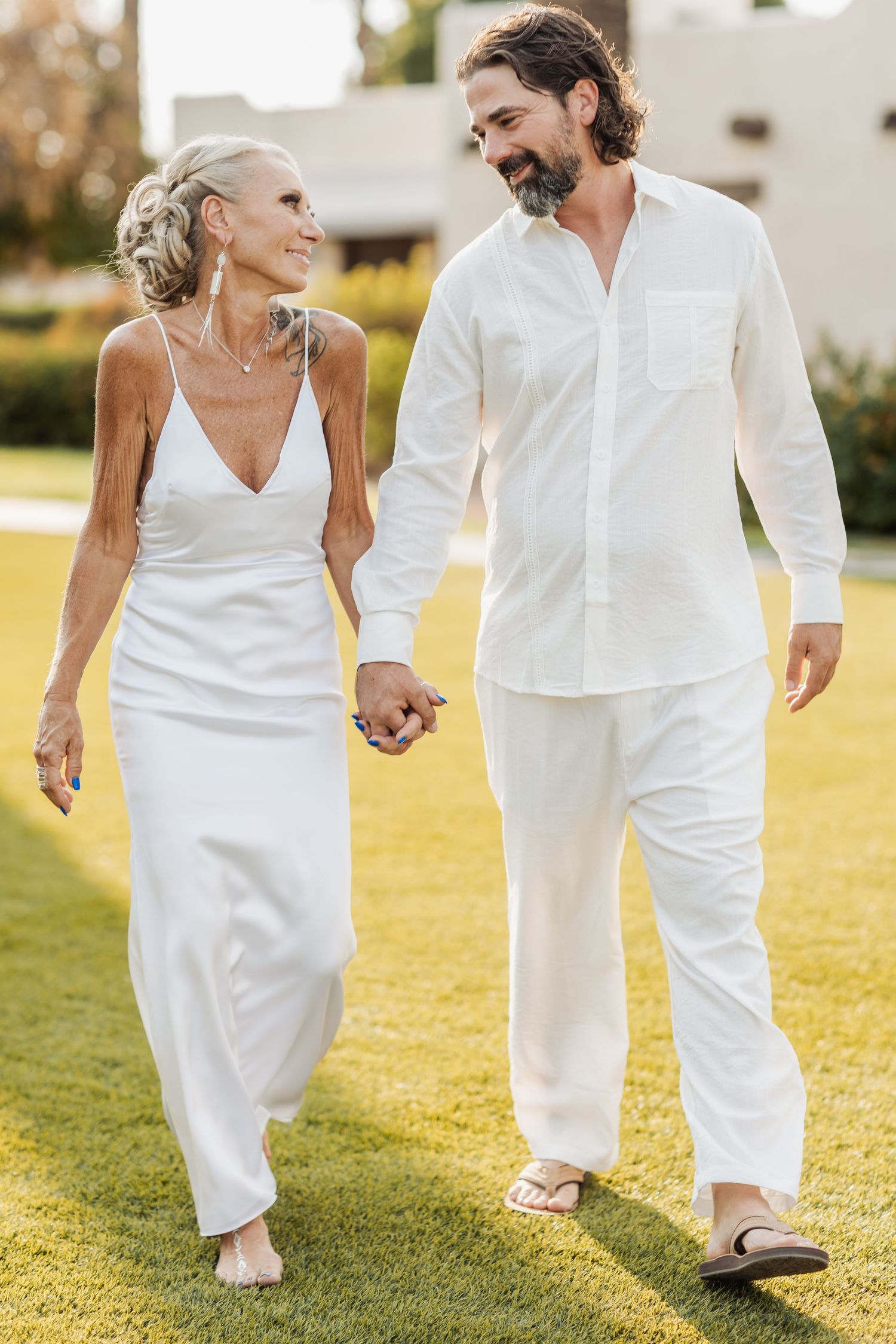 Couple in all-white attire walking hand in hand on sun-drenched grass during golden hour.