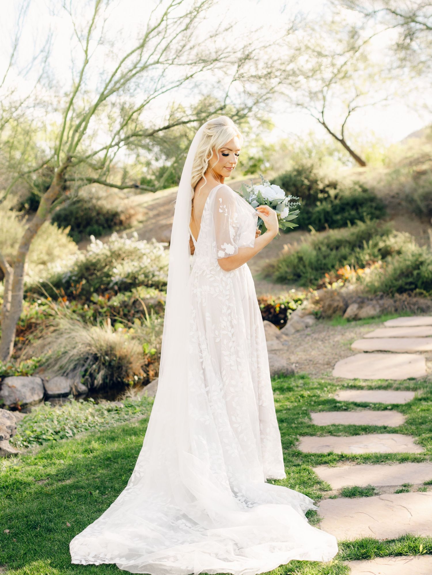 A bride in a flowing white gown walks along a stone path surrounded by desert landscaping on her wedding day.