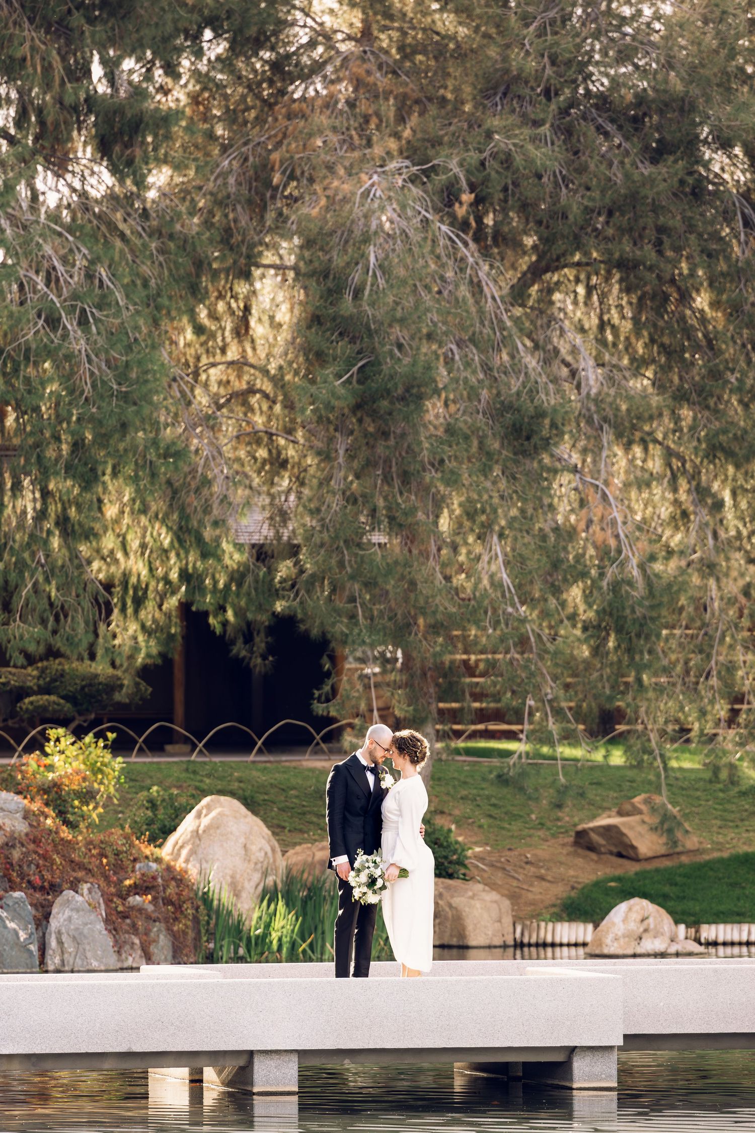 A romantic wedding couple stands on a white dock by a tranquil pond surrounded by lush greenery at sunset.
