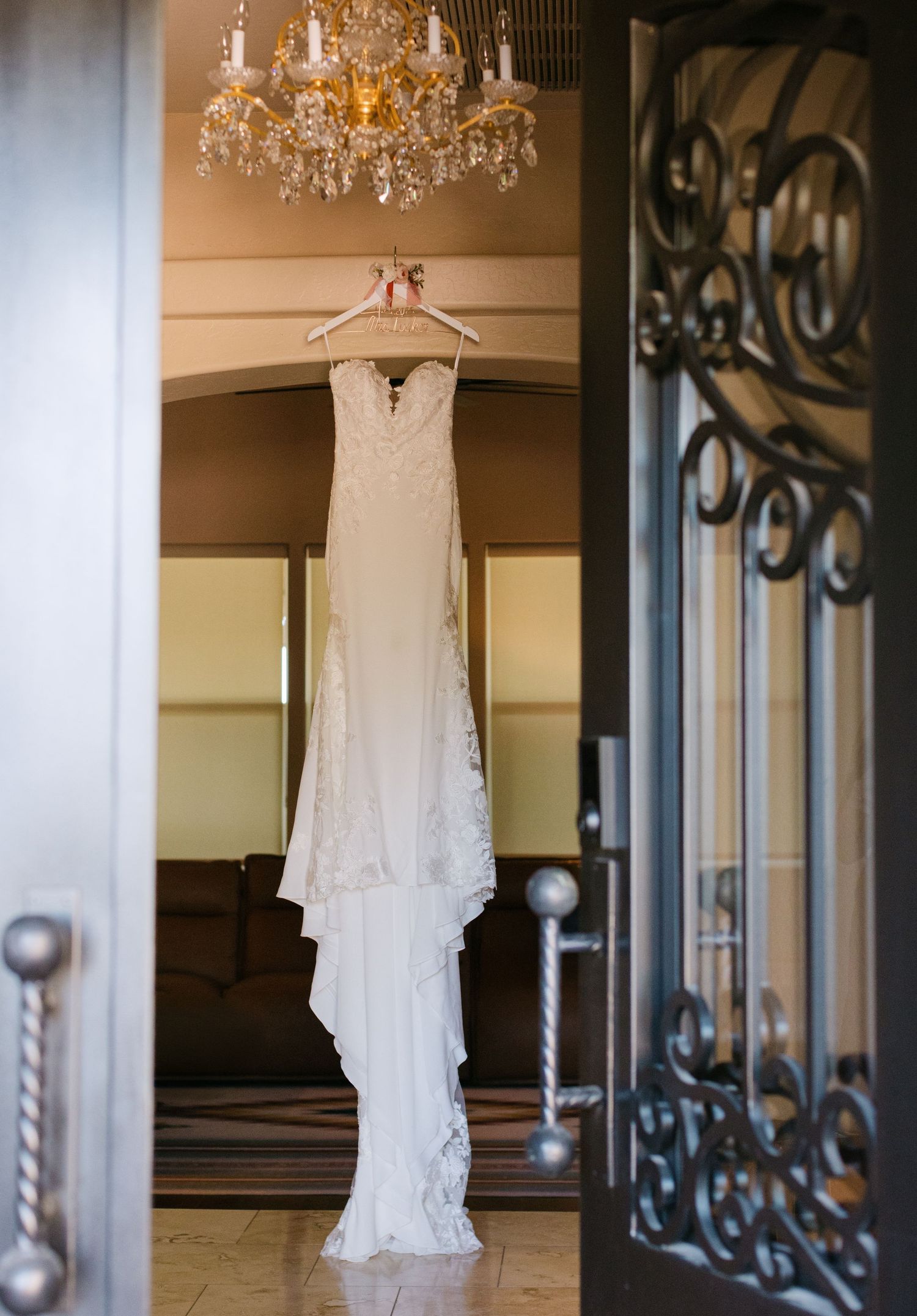 A delicate lace wedding dress hangs elegantly from a crystal chandelier in an ornate room with wrought iron details.