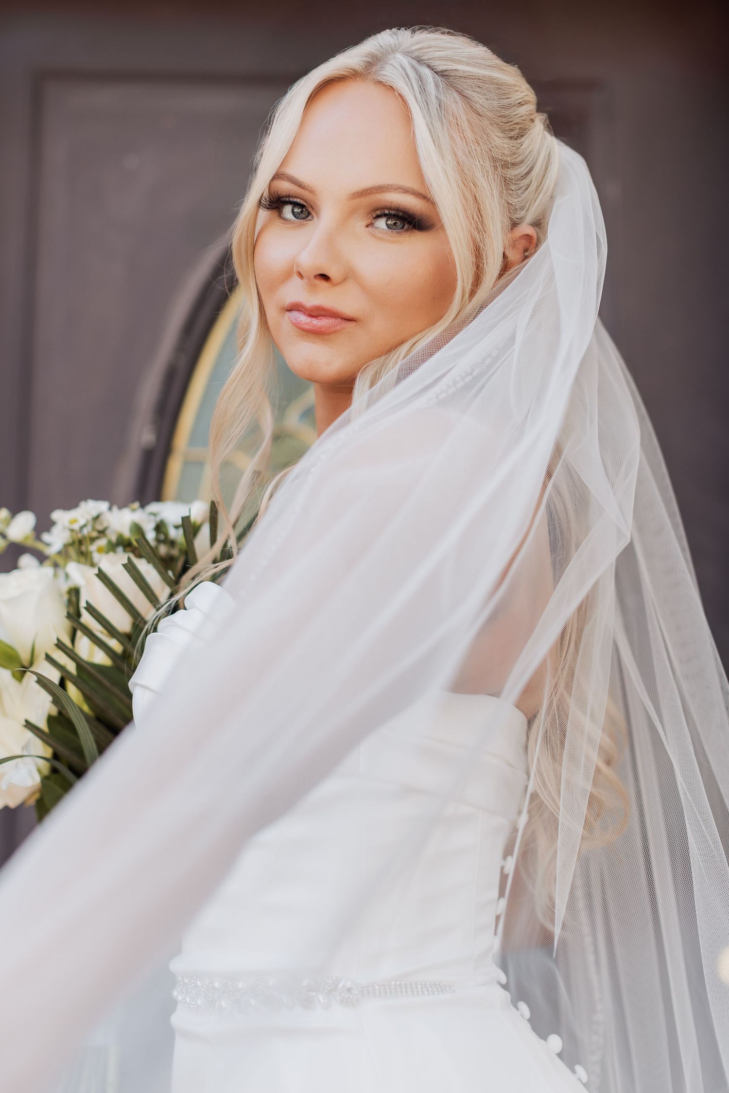 A bride in an elegant white wedding dress and flowing veil holds a bouquet while posing on her wedding day.