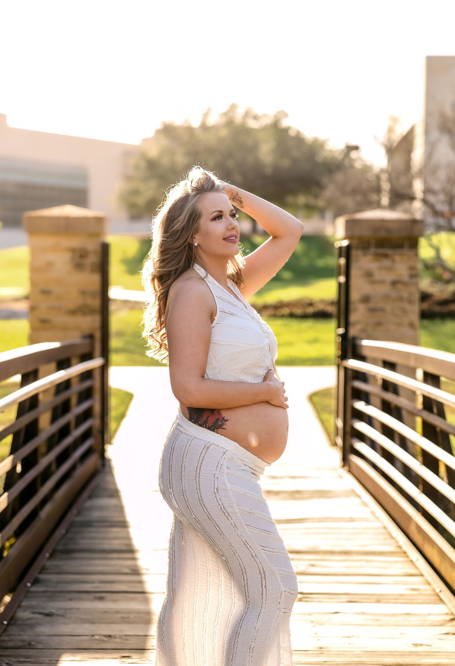 Maternity photo of an expectant mother in white attire posing on a wooden bridge at sunset.