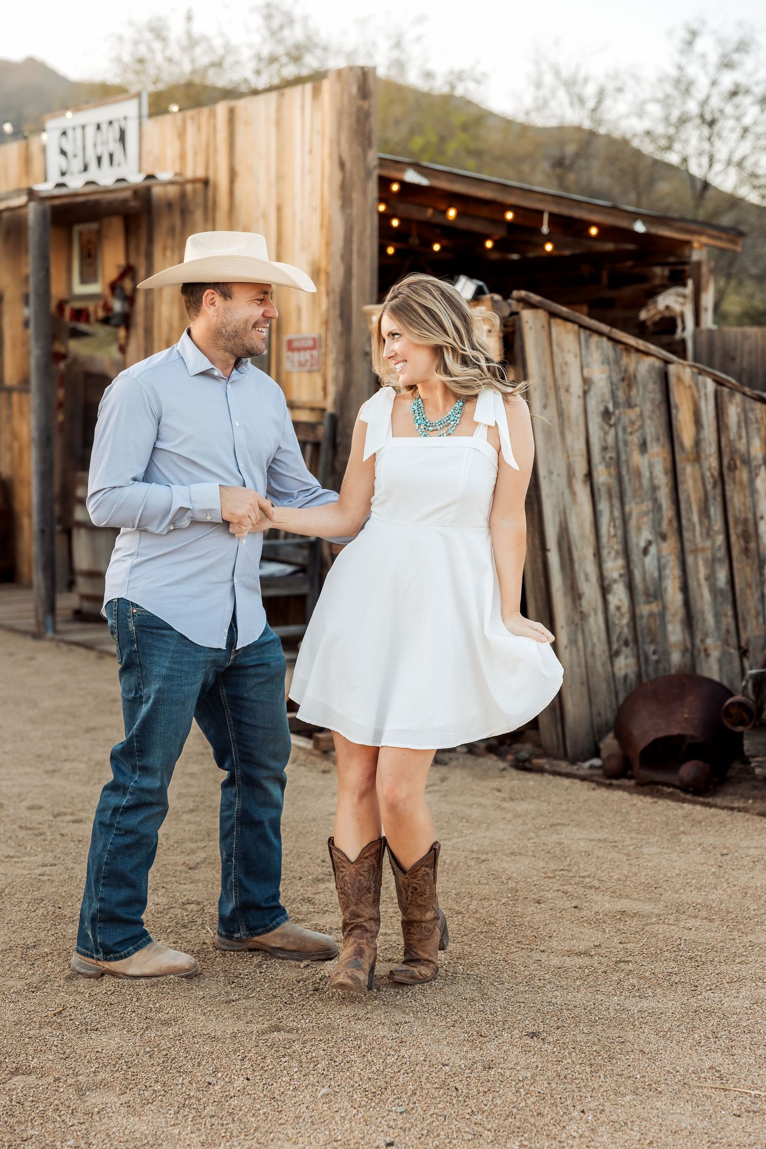 Couple dancing together in a rustic outdoor setting with wooden structures and dirt ground.