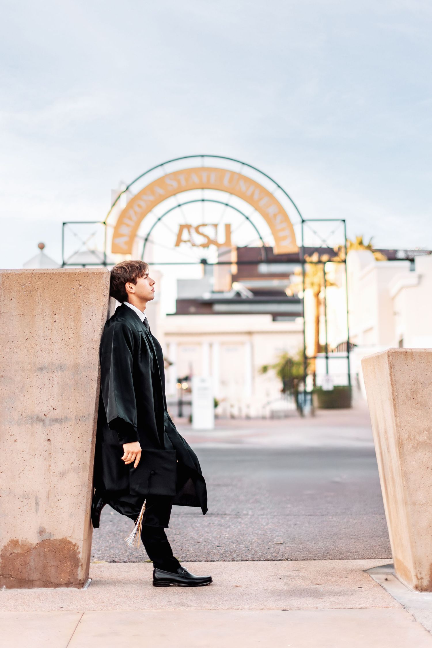 A graduate stands against a concrete wall near an ASU campus archway entrance during a graduation photoshoot.