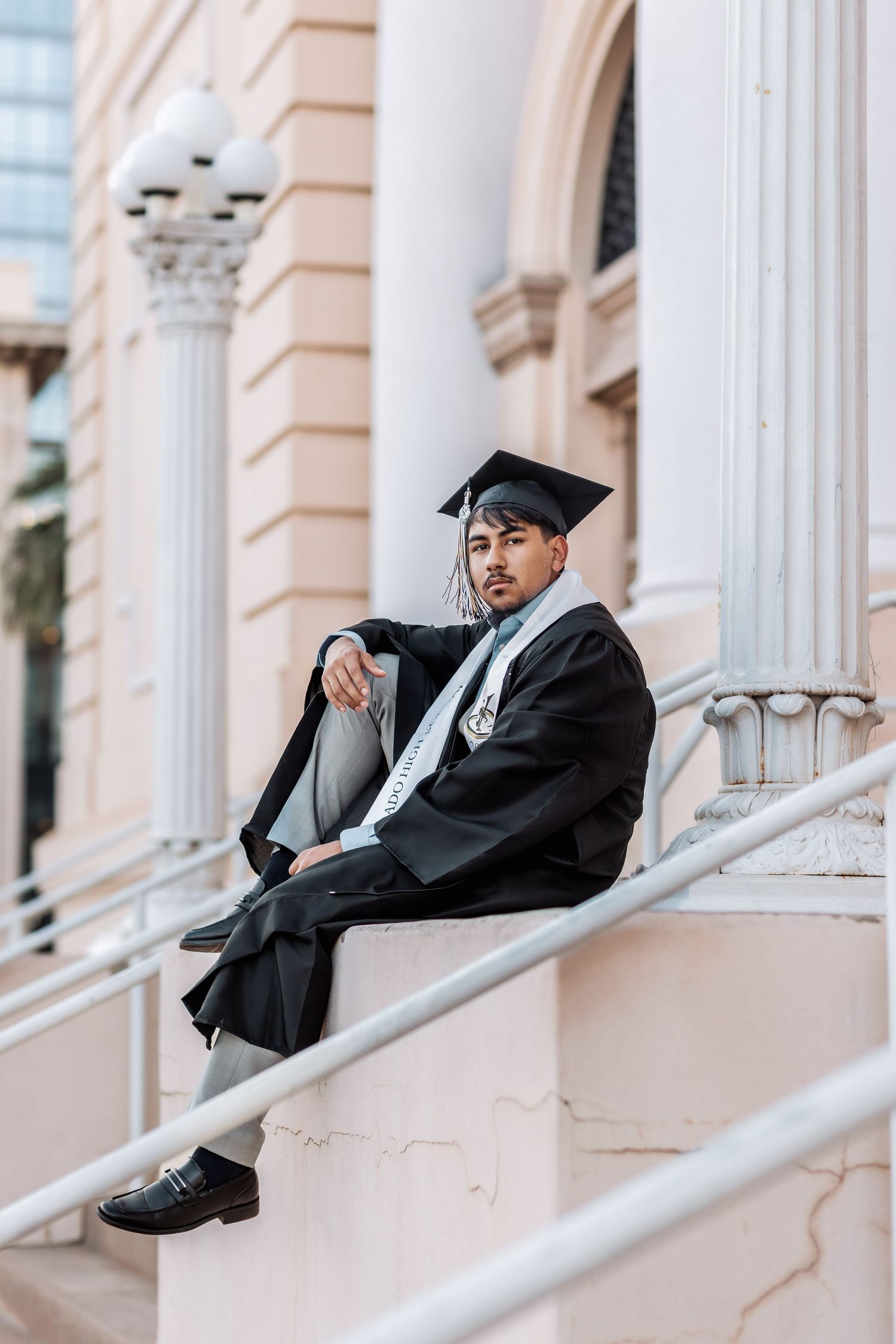 Graduate in cap and gown sits pensively on white marble steps of classical building with ornate columns.