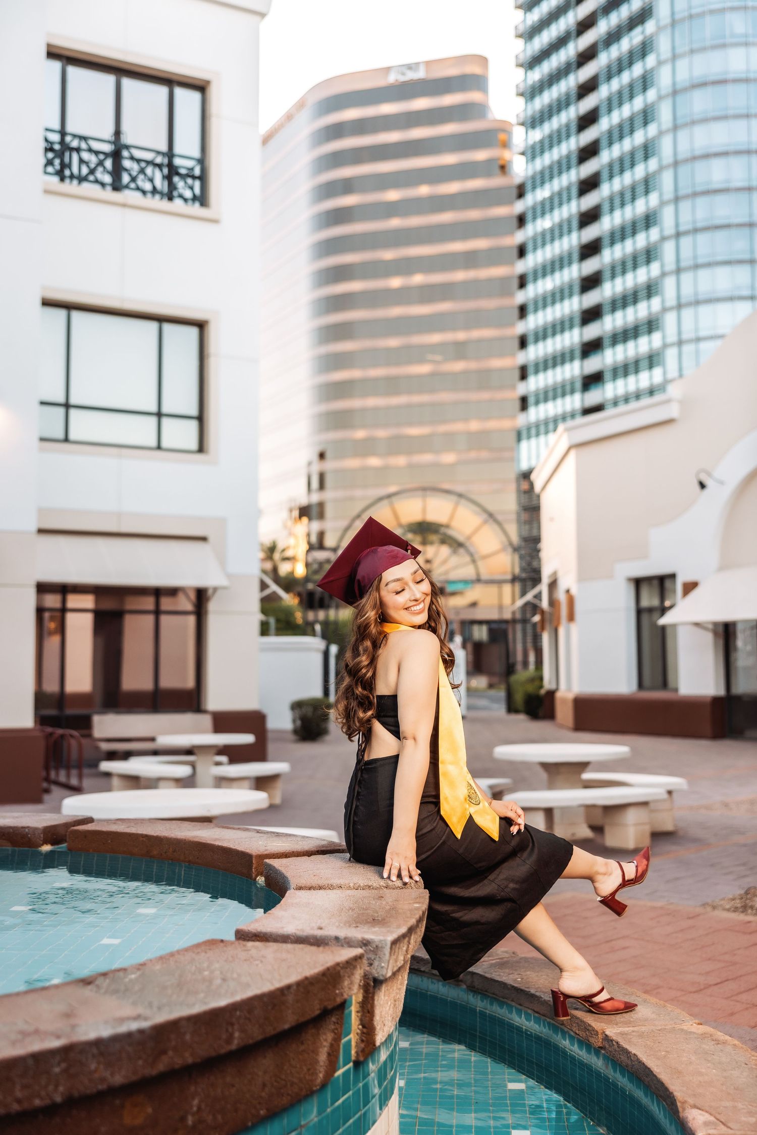 A graduate in a black dress and yellow stole sits by a fountain with modern city buildings in the background.
