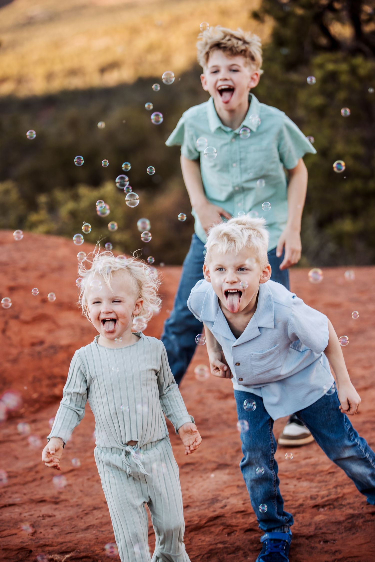 Children playing and laughing while chasing soap bubbles on red clay ground in a playful outdoor scene.