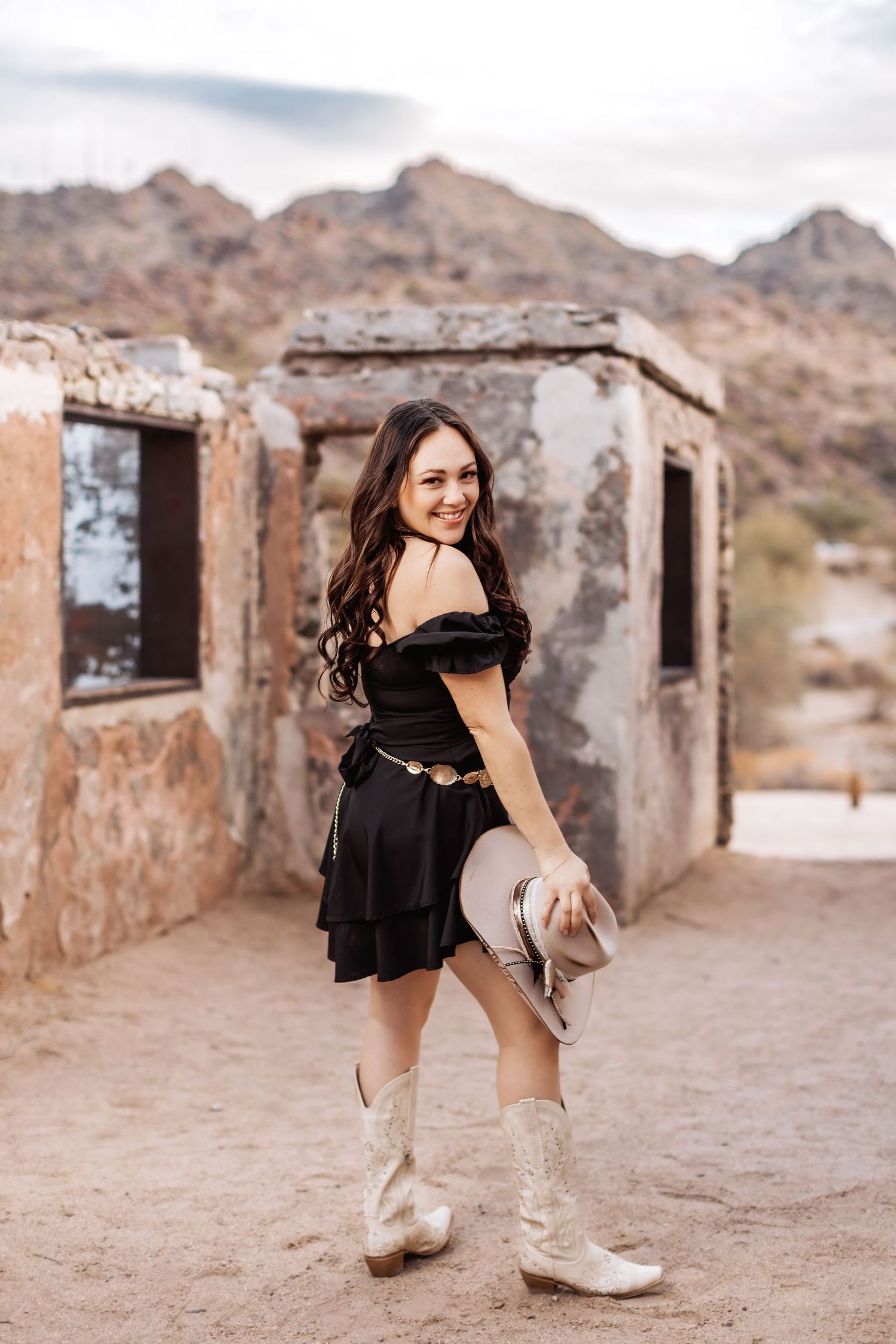 A person in a black dress and cowboy boots poses in front of weathered stone ruins in a desert landscape.
