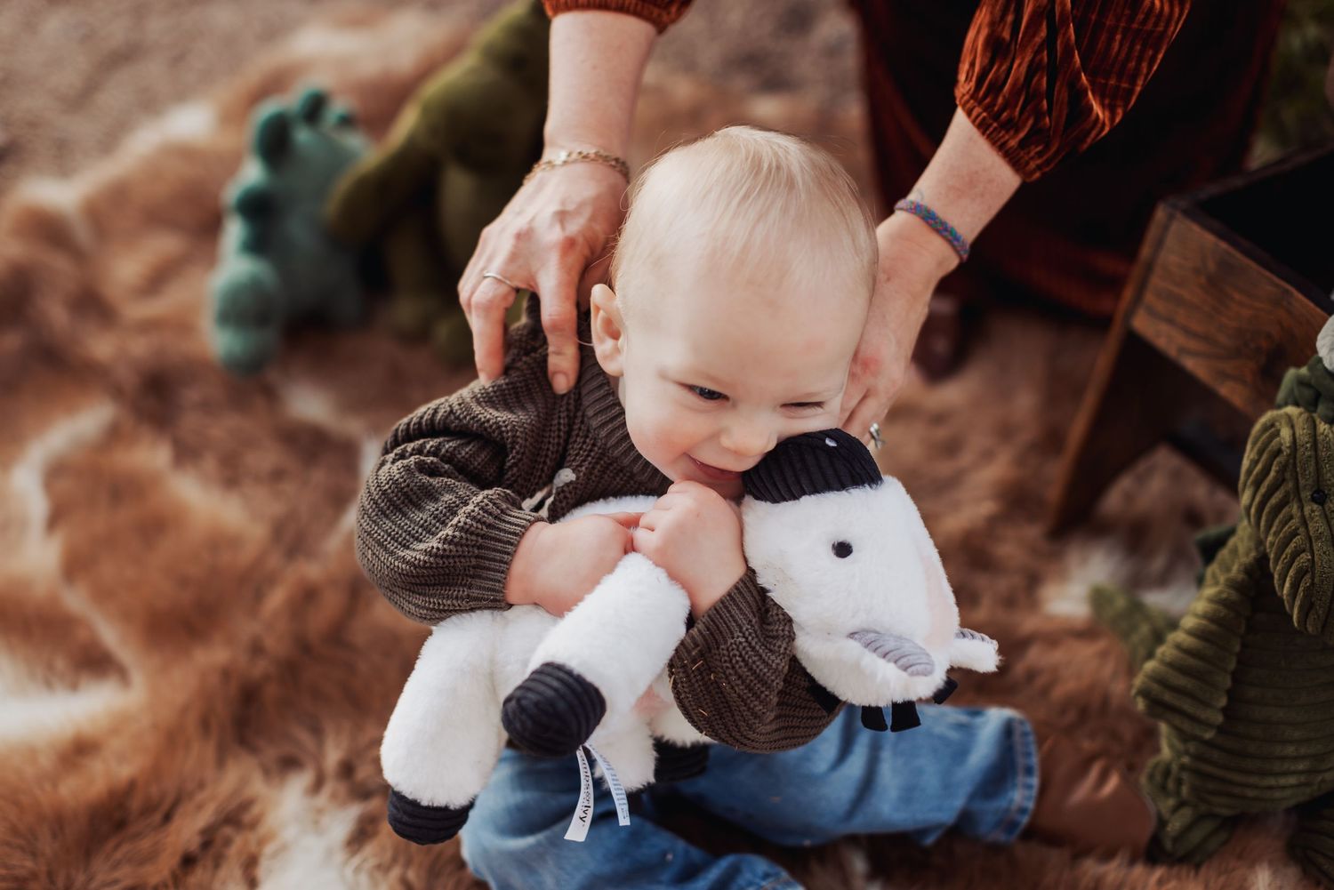 A small baby holds a plush lamb toy while sitting on a brown textured blanket with a caring hand offering support.