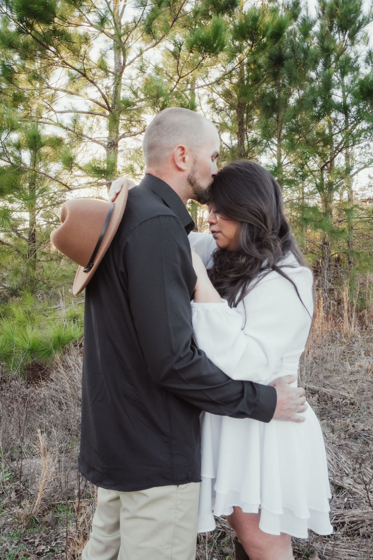 A romantic outdoor engagement photo in a pine forest with a couple embracing in casual white and black attire.