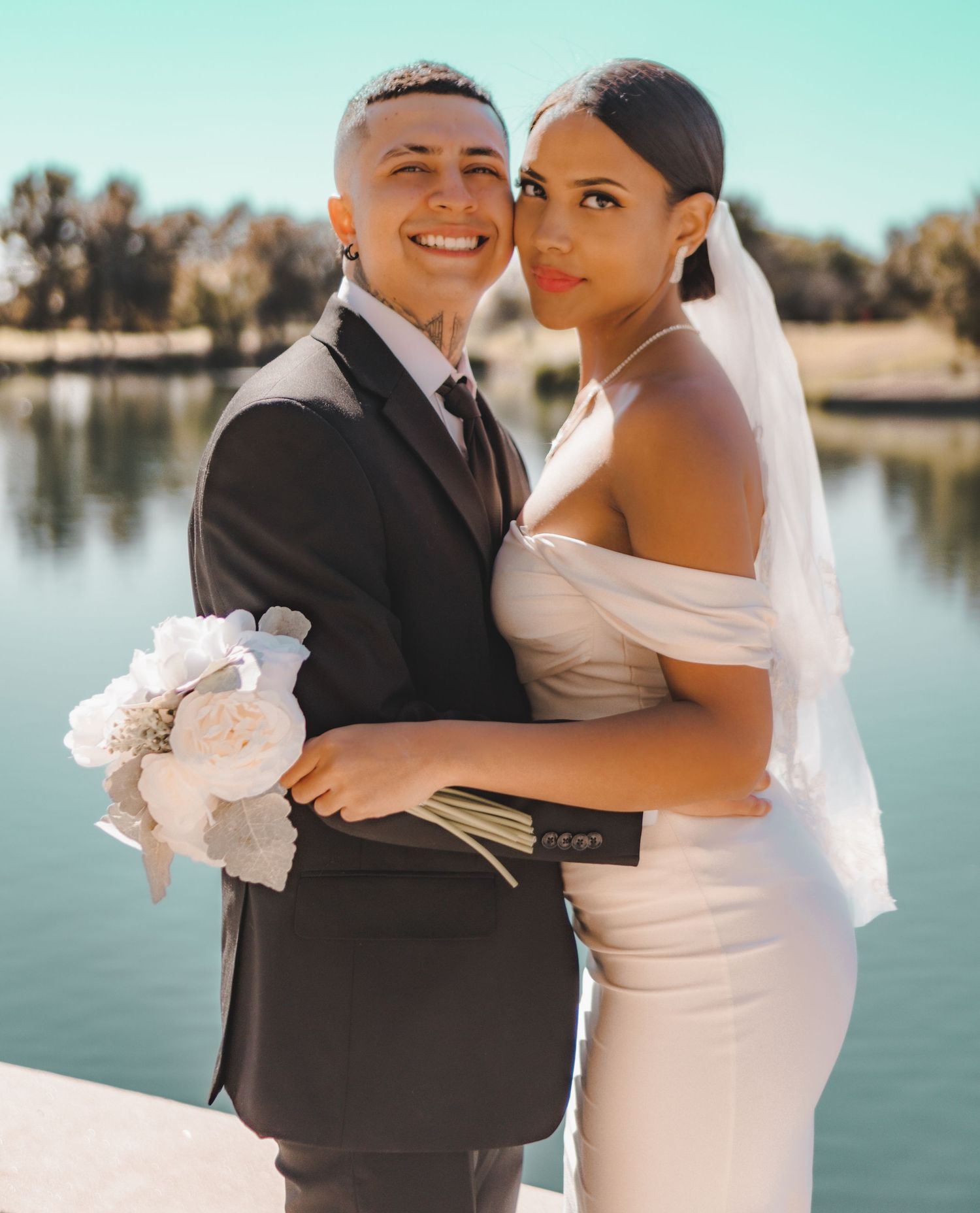 A bride and groom pose for their wedding photo by a lake on a sunny day.