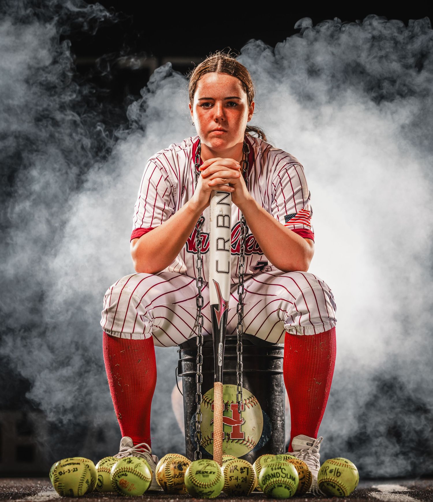 Female softball player in pinstripe uniform sits on bat surrounded by softballs in dramatic smoky setting.