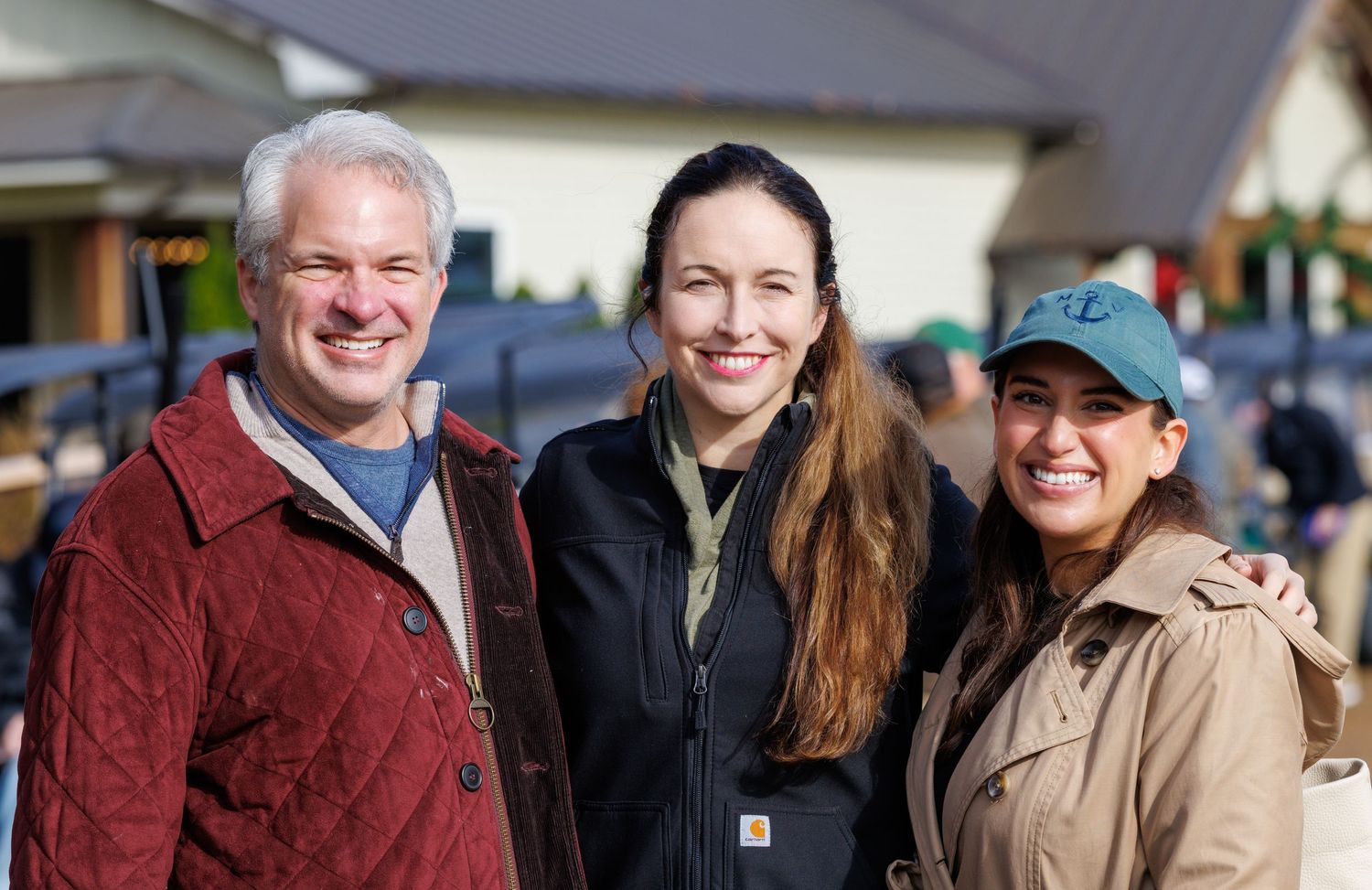 Three smiling people posing together outdoors at a casual event on a sunny day.
