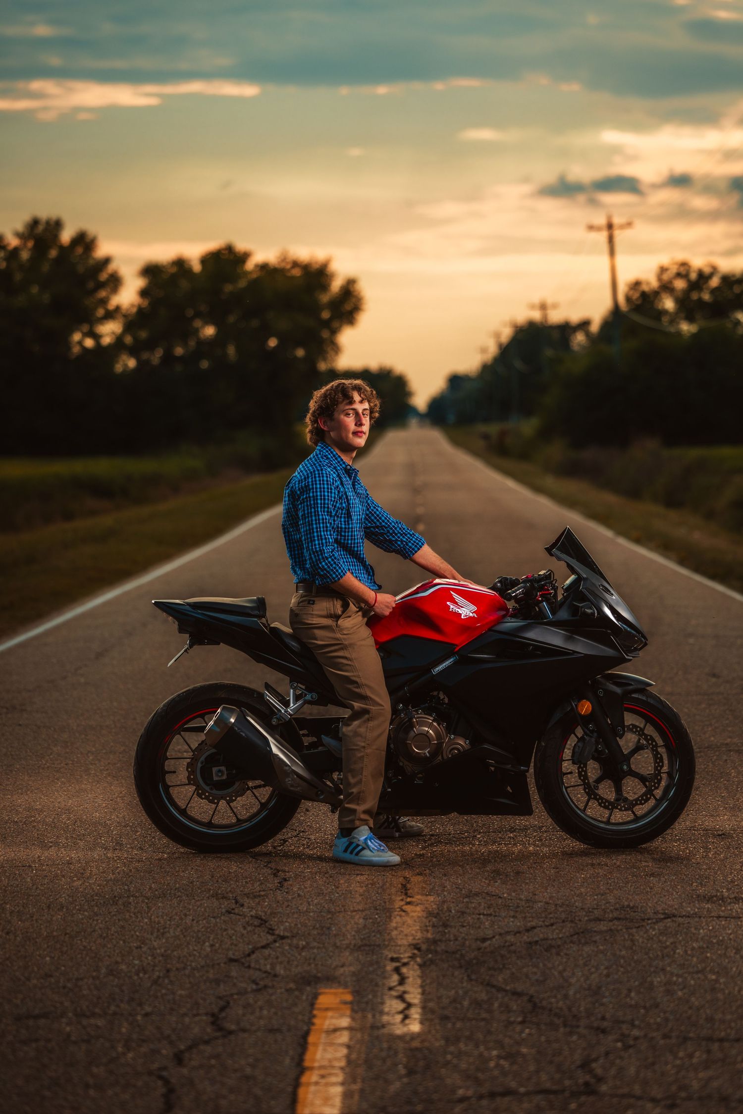 Young man posing with red and black sport motorcycle on empty road at dramatic sunset.