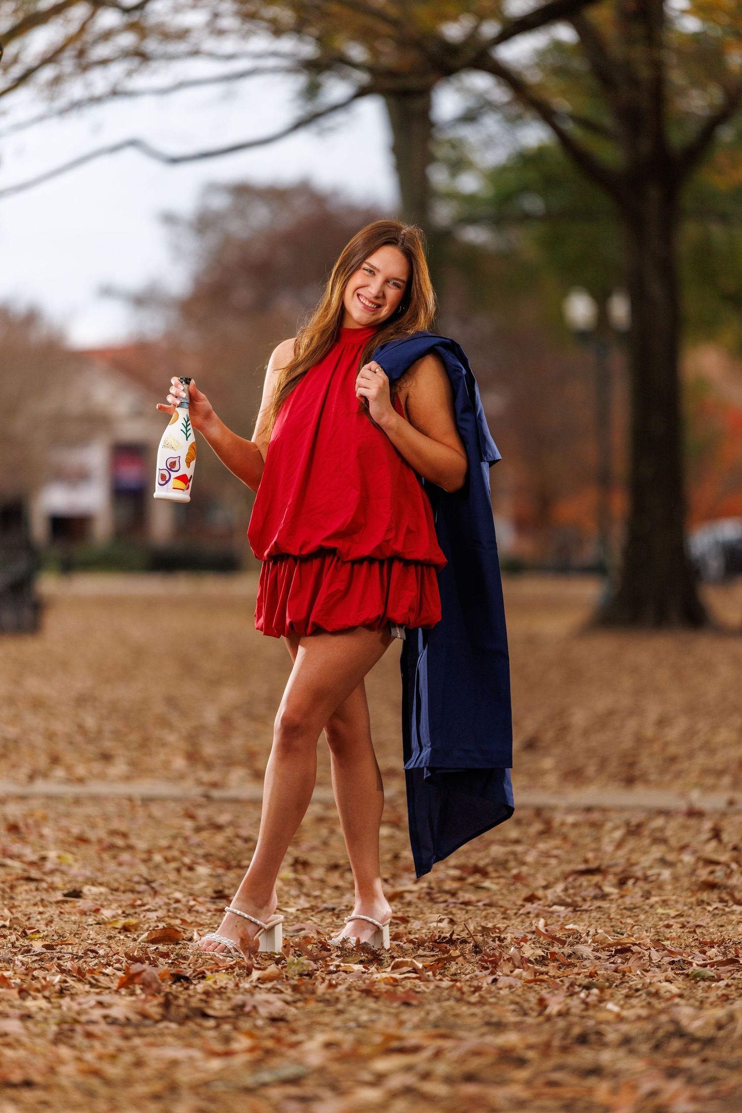 Smiling woman in red dress holding decorated bottle with blue jacket over shoulder in autumn park.