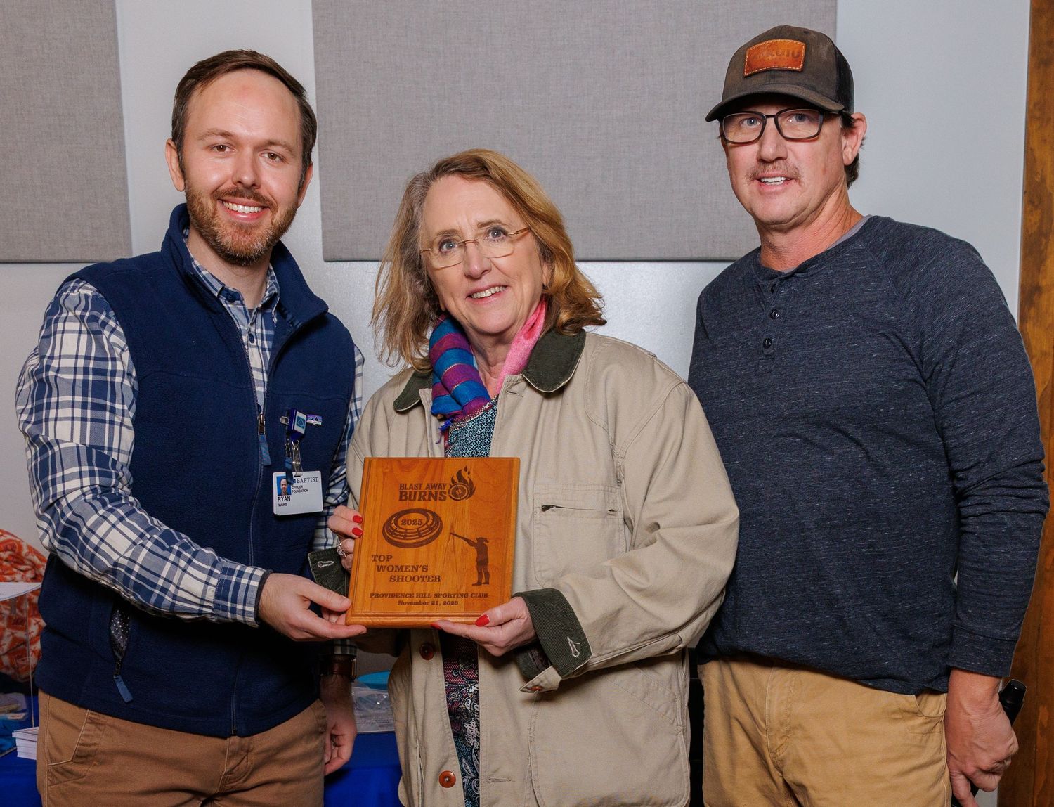 Three people smiling while holding a wooden award plaque at an indoor recognition event.
