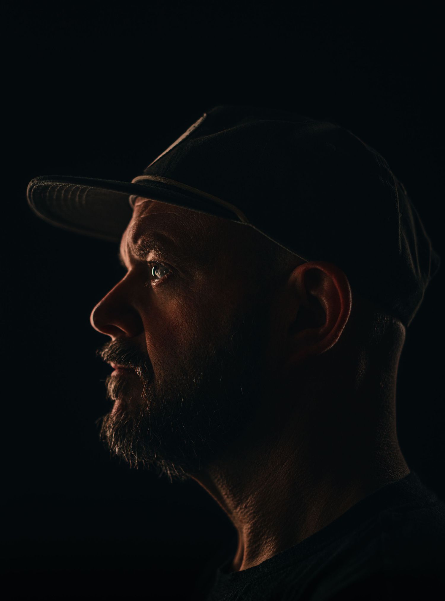 Dramatic side profile portrait of bearded man wearing baseball cap against dark background.