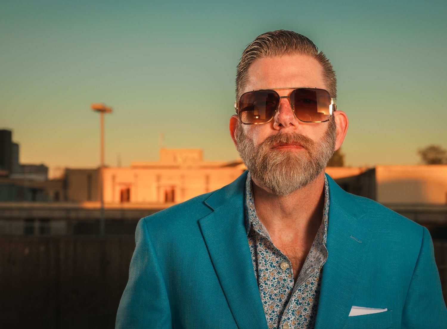 Confident bearded man in teal blazer and sunglasses posing outdoors at golden hour.