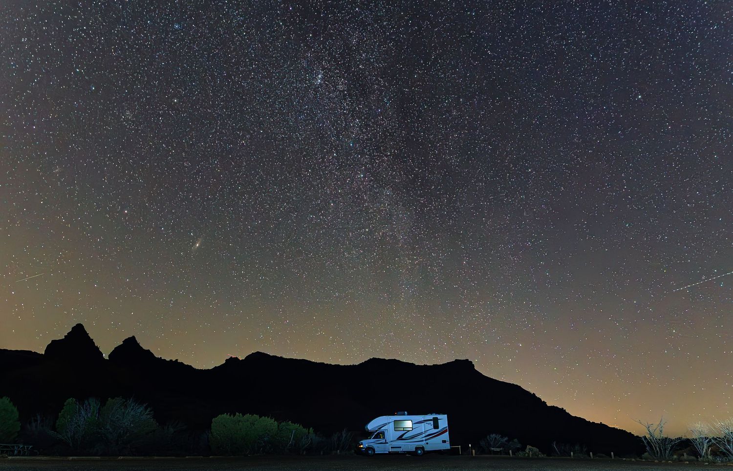 RV parked under stunning Milky Way galaxy and starry night sky near mountain silhouette.