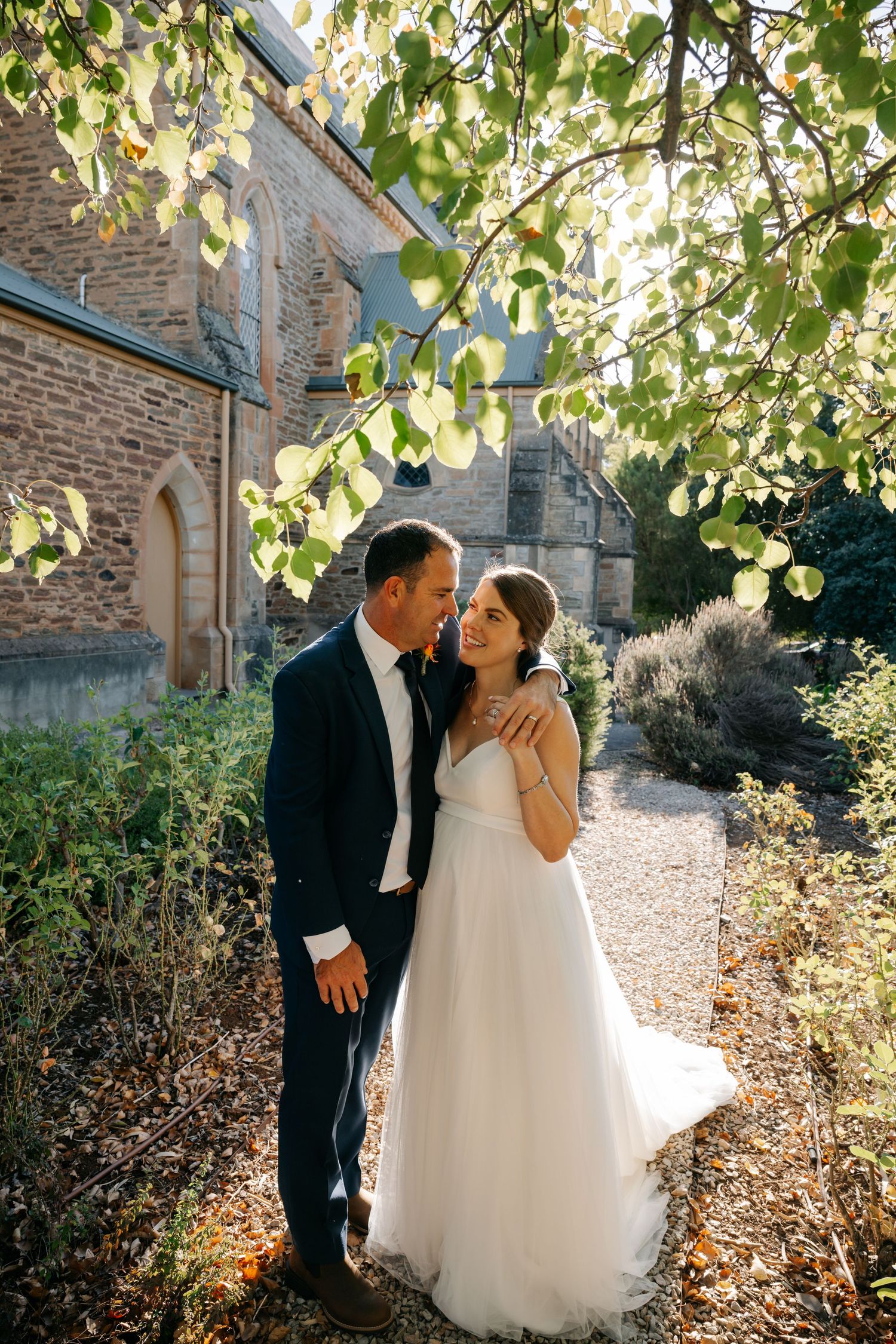 A romantic wedding couple shares an intimate moment in the sunlit garden of a stone church.