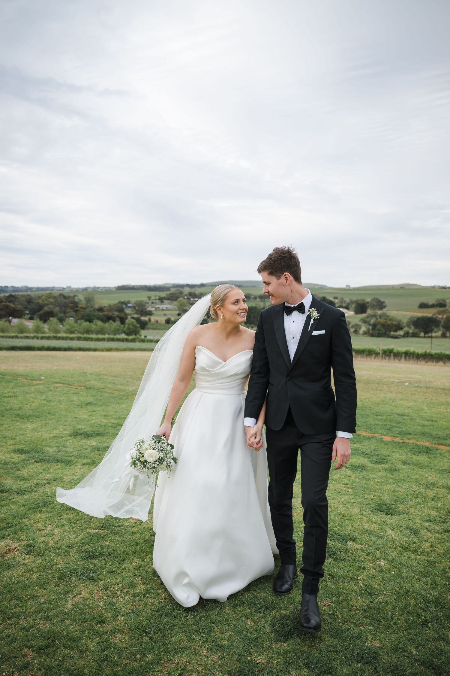 A couple in wedding attire walks together across a scenic green countryside field on their special day.